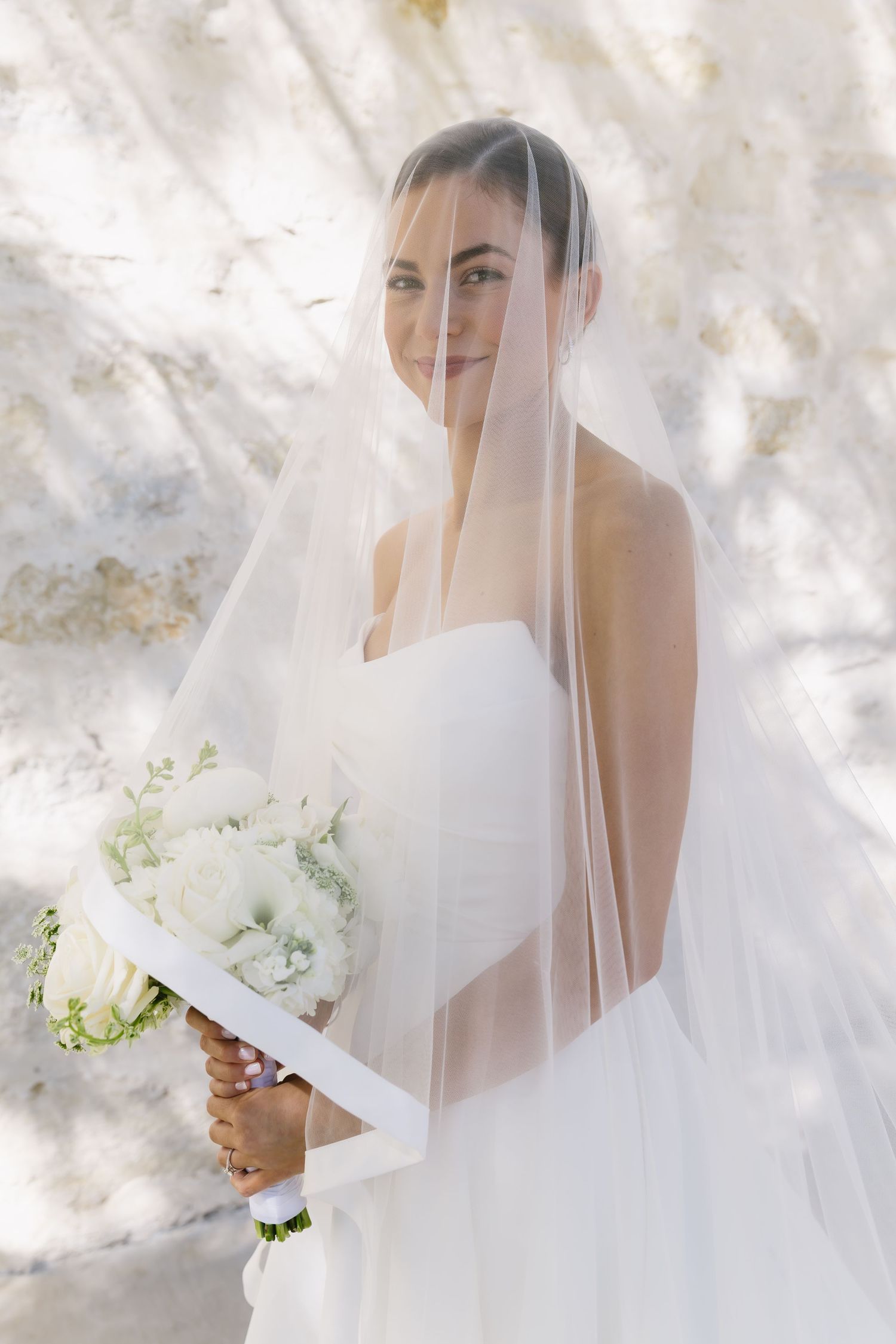 Ethereal bridal portrait with delicate veil and white bouquet against a sunlit backdrop.