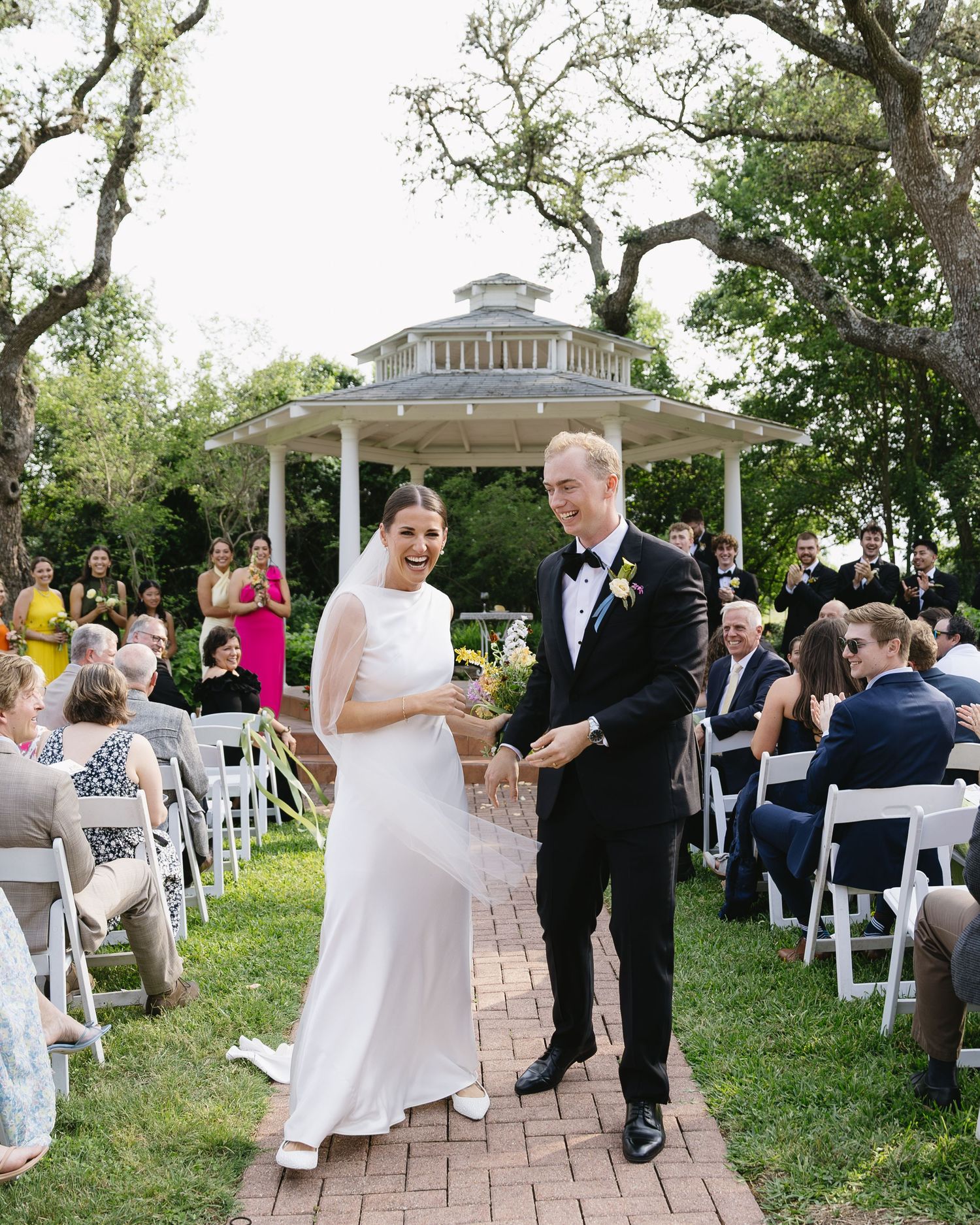 Outdoor wedding ceremony with guests seated on white chairs under trees on a brick patio.