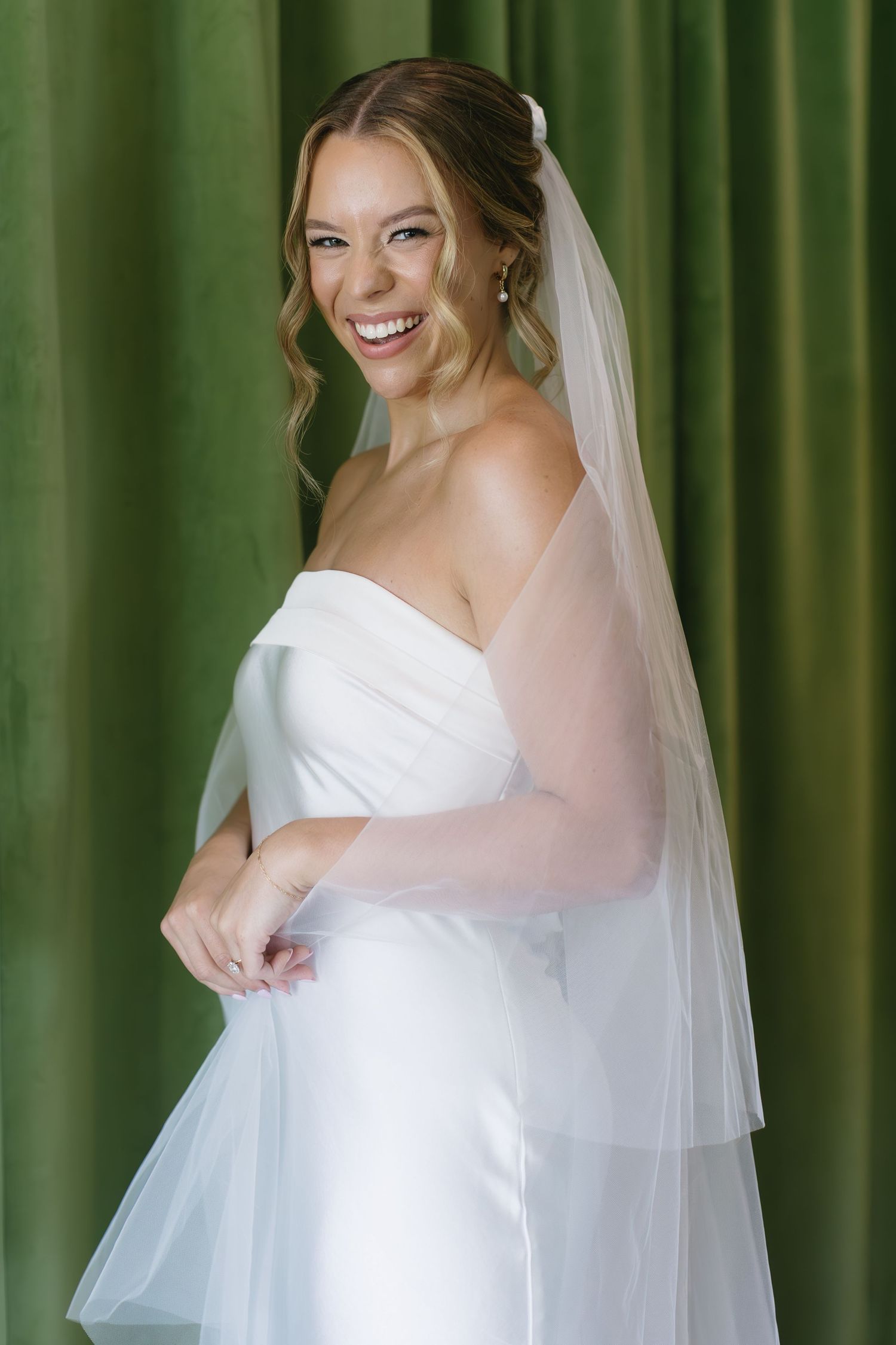 Elegant bride in white gown smiles while standing against green curtain backdrop.
