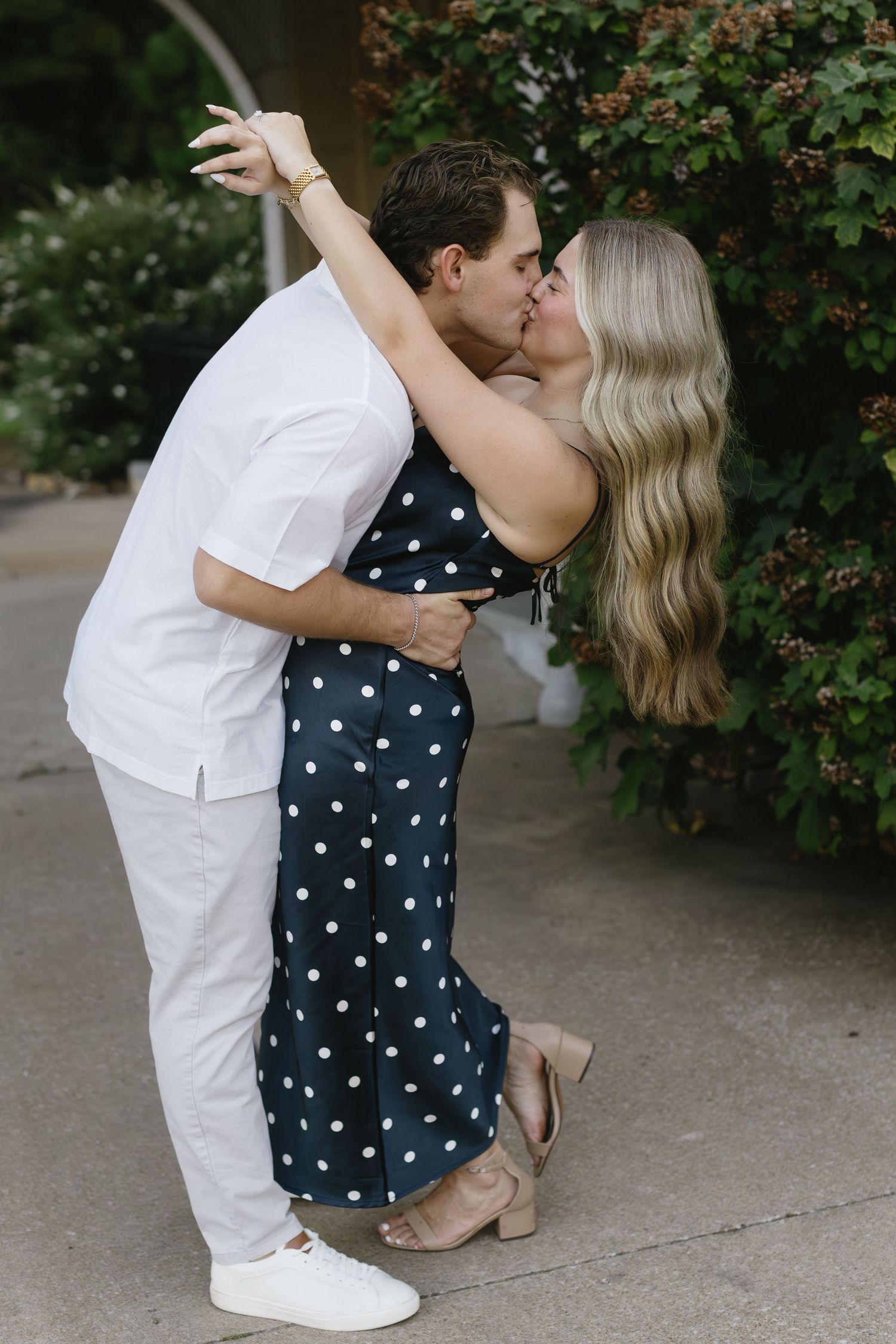 A romantic couple shares a kiss outdoors, with one wearing a polka dot dress and the other in white attire.