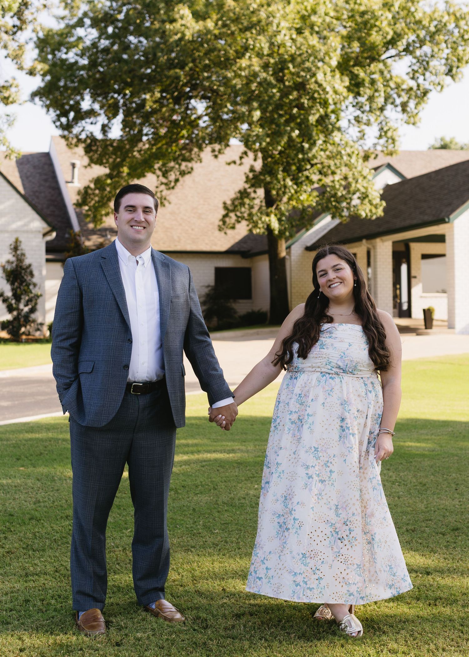 A couple poses together outdoors, with the woman wearing a floral dress and the man in a navy suit.