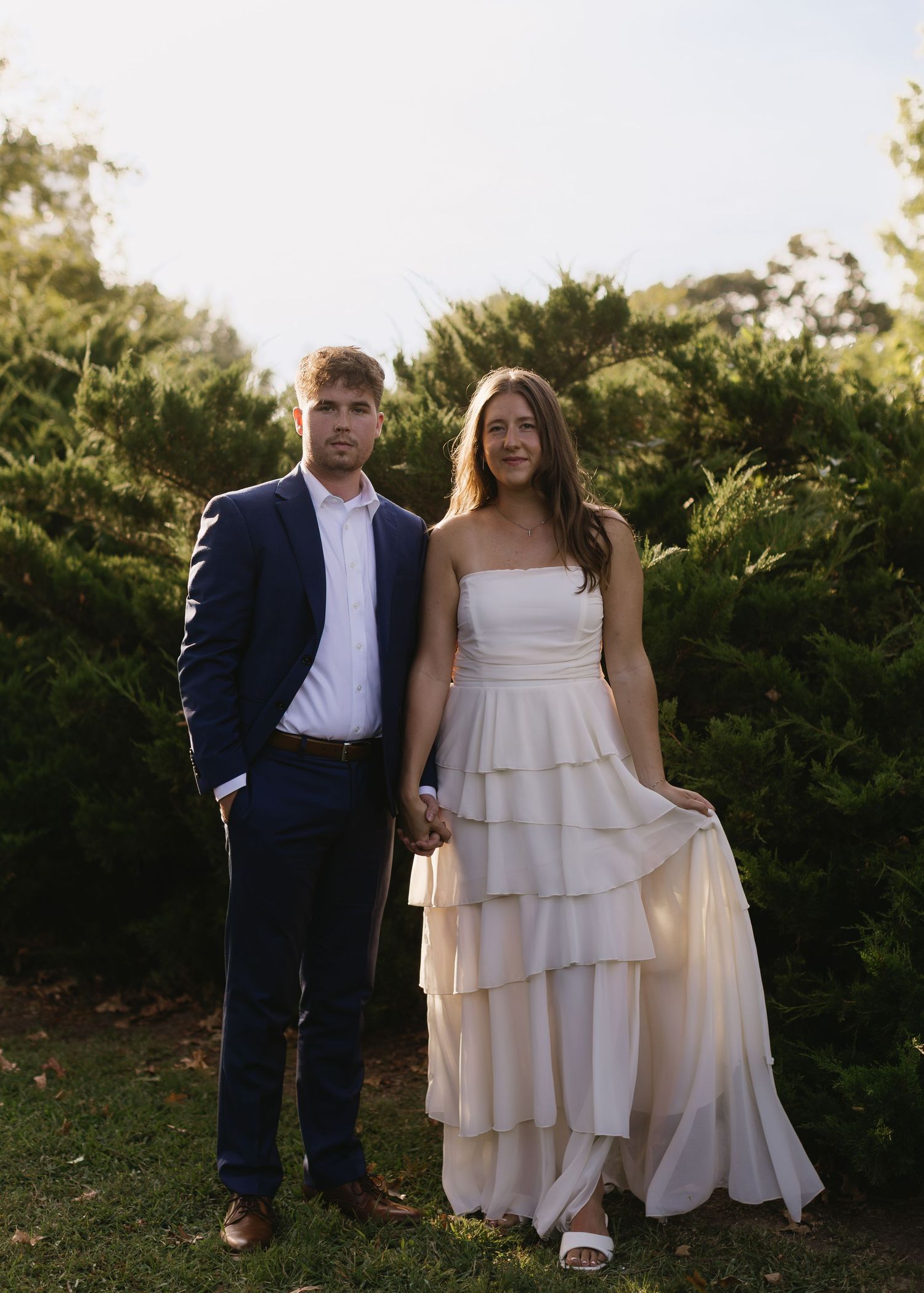A couple in formal attire poses together on a grassy lawn with greenery in the background.