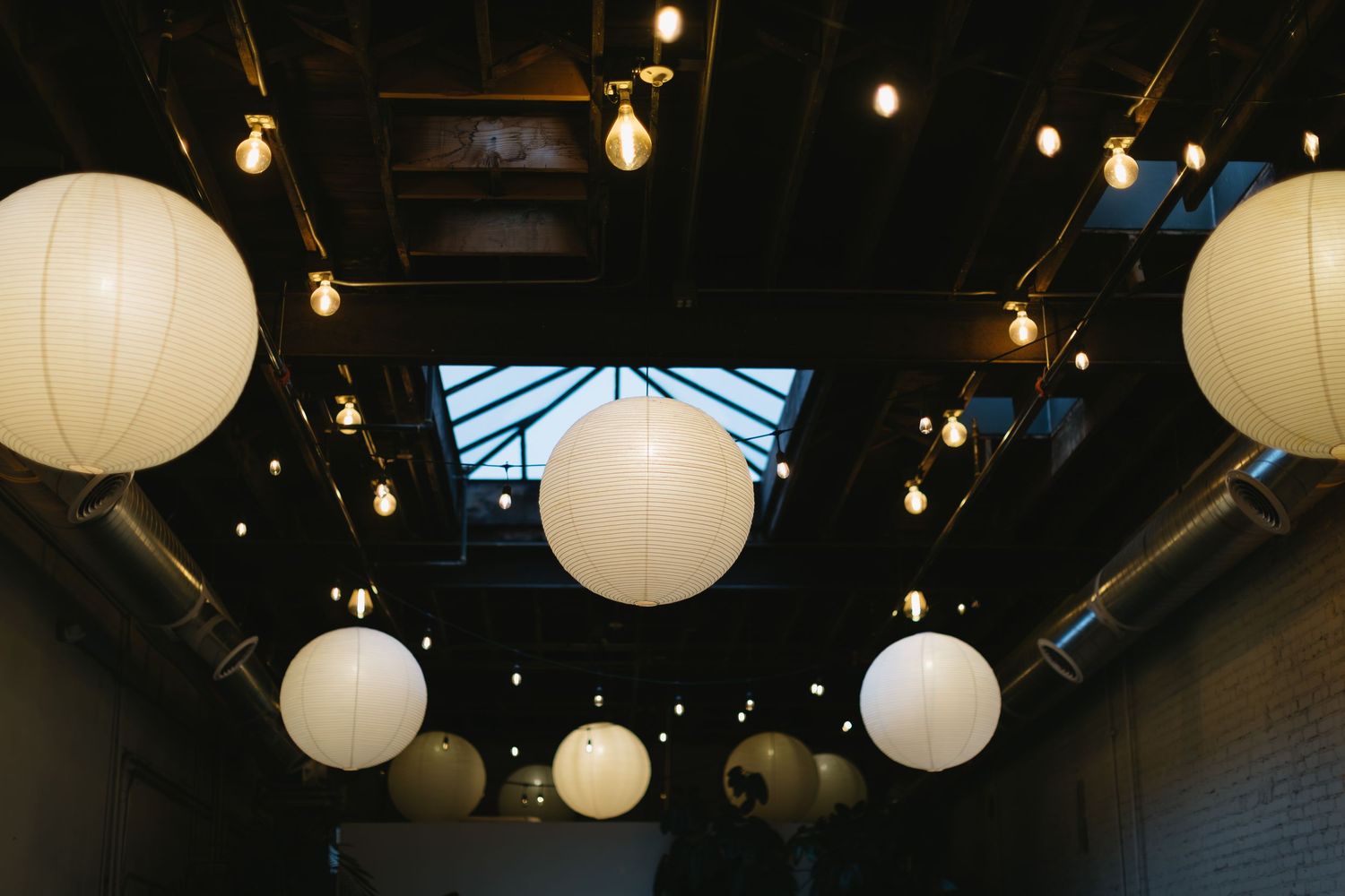 String lights and white paper lanterns hanging from a dark ceiling create ambient lighting.