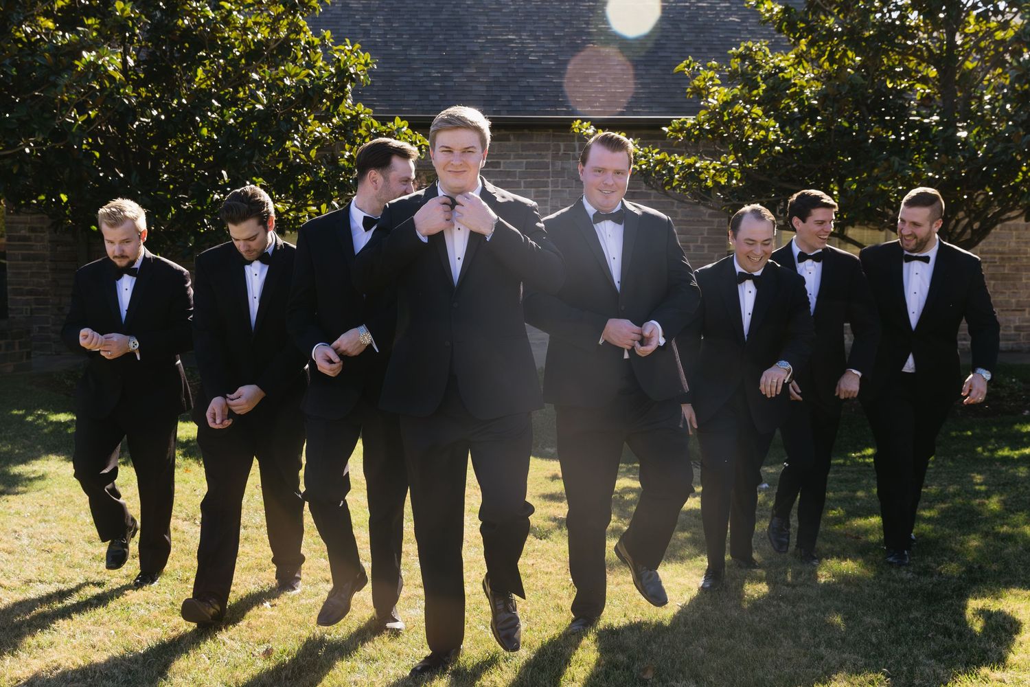 Wedding party in black formal attire poses outdoors with trees in background.