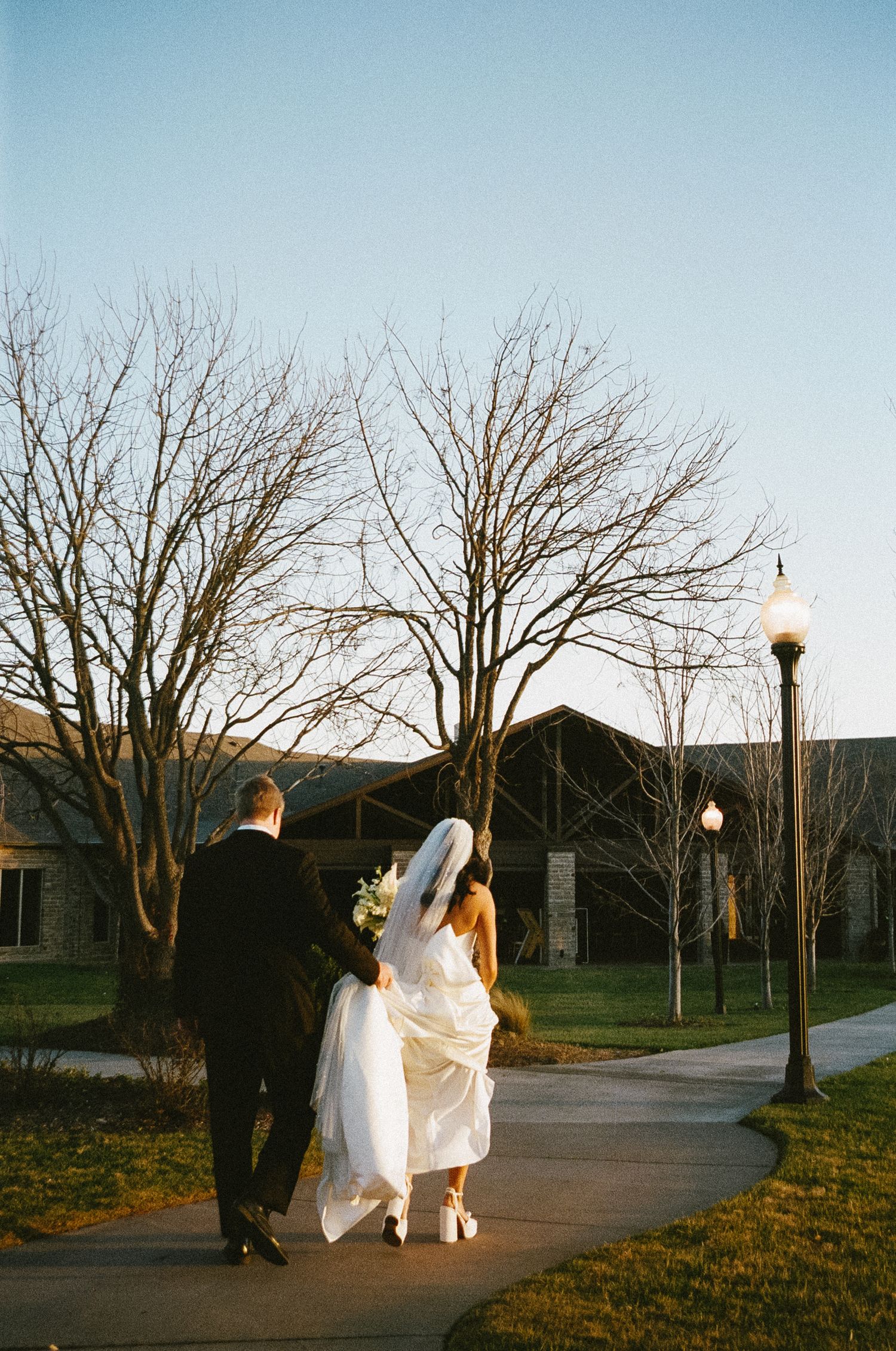 Wedding couple walking along tree-lined path at sunset with flowing white dress.