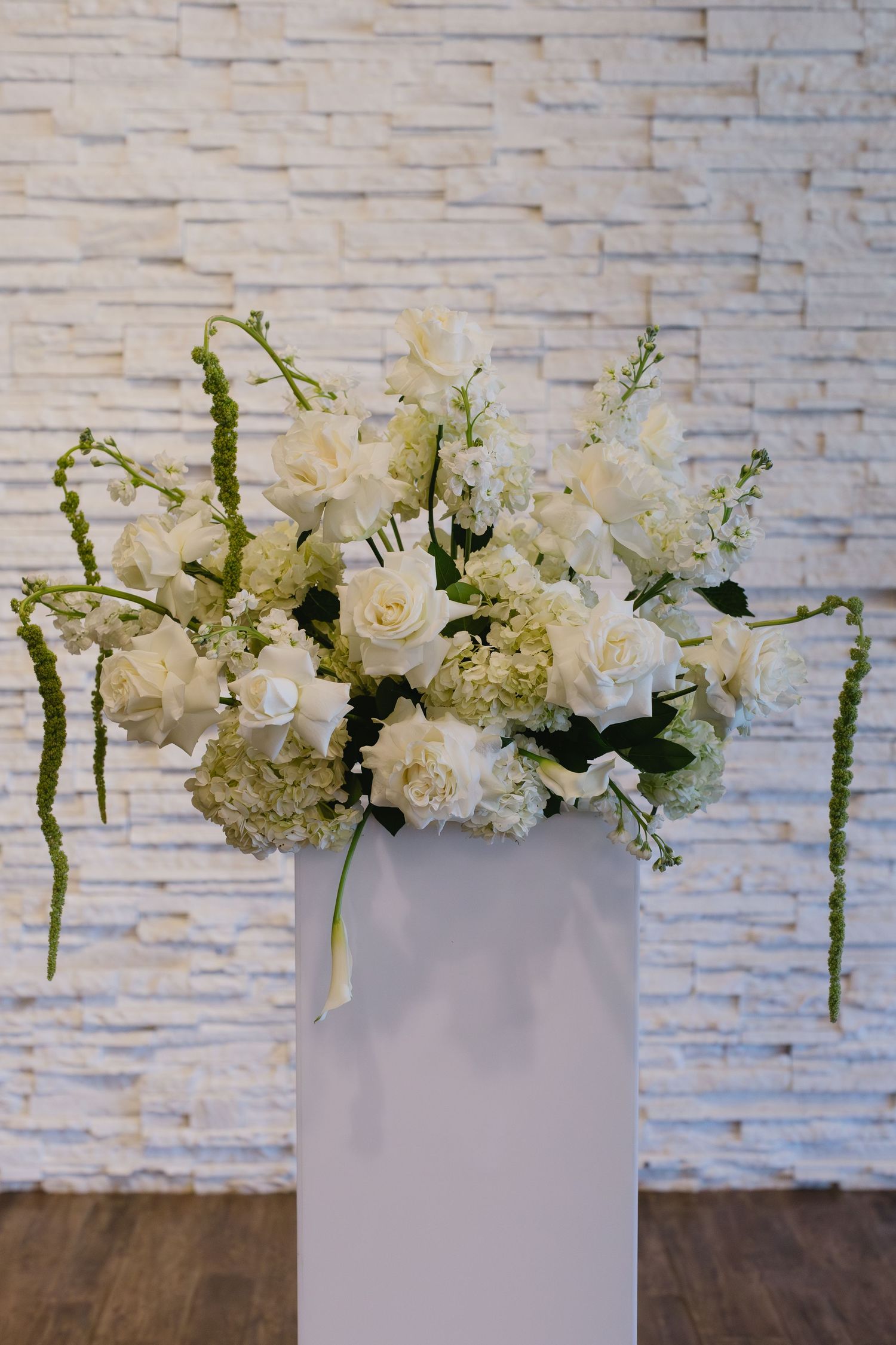 Elegant white floral arrangement with roses and hanging amaranthus in modern white vase against textured wall.