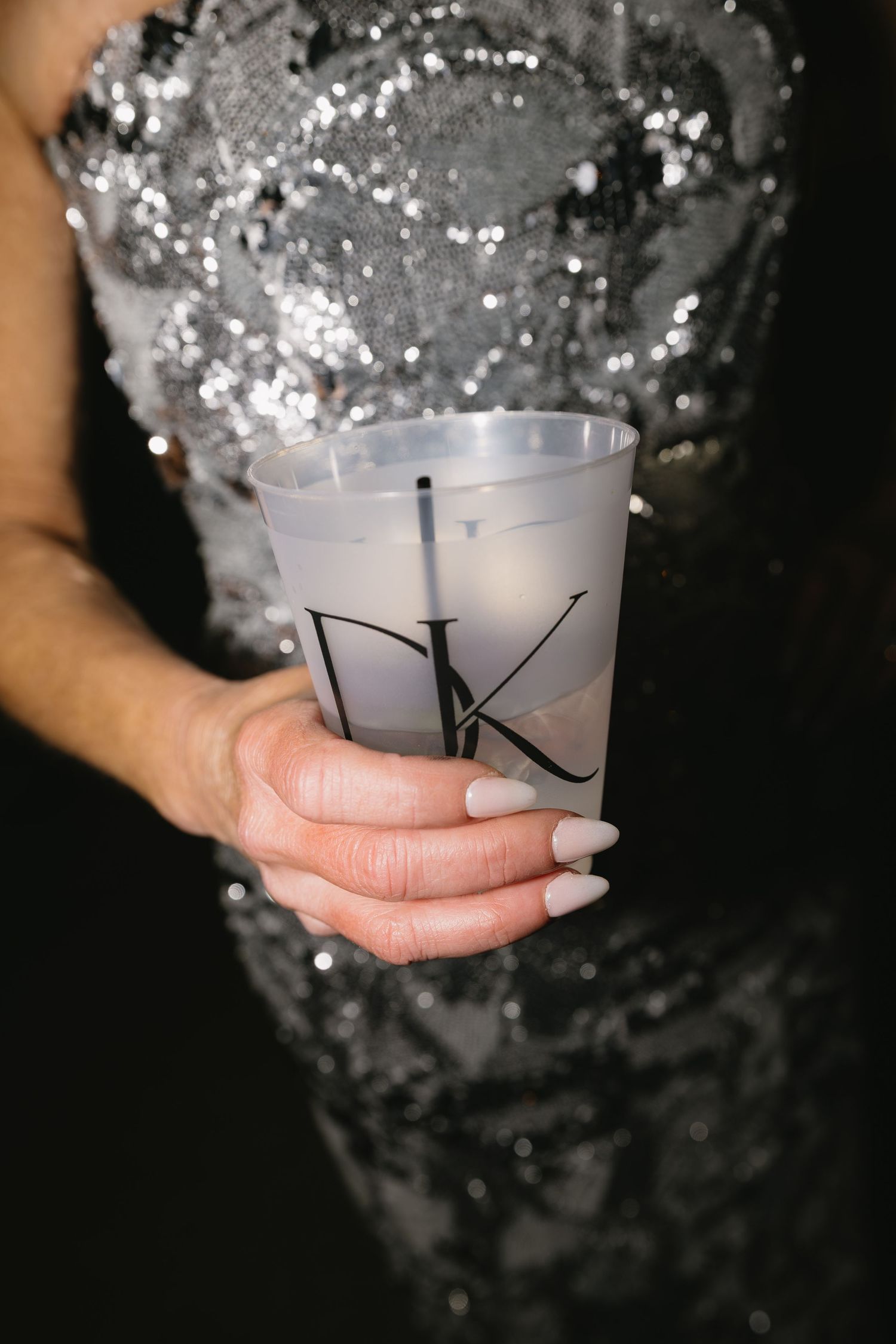 A hand holding a frosted drink cup with monogrammed DK initials against a sparkly dress background.
