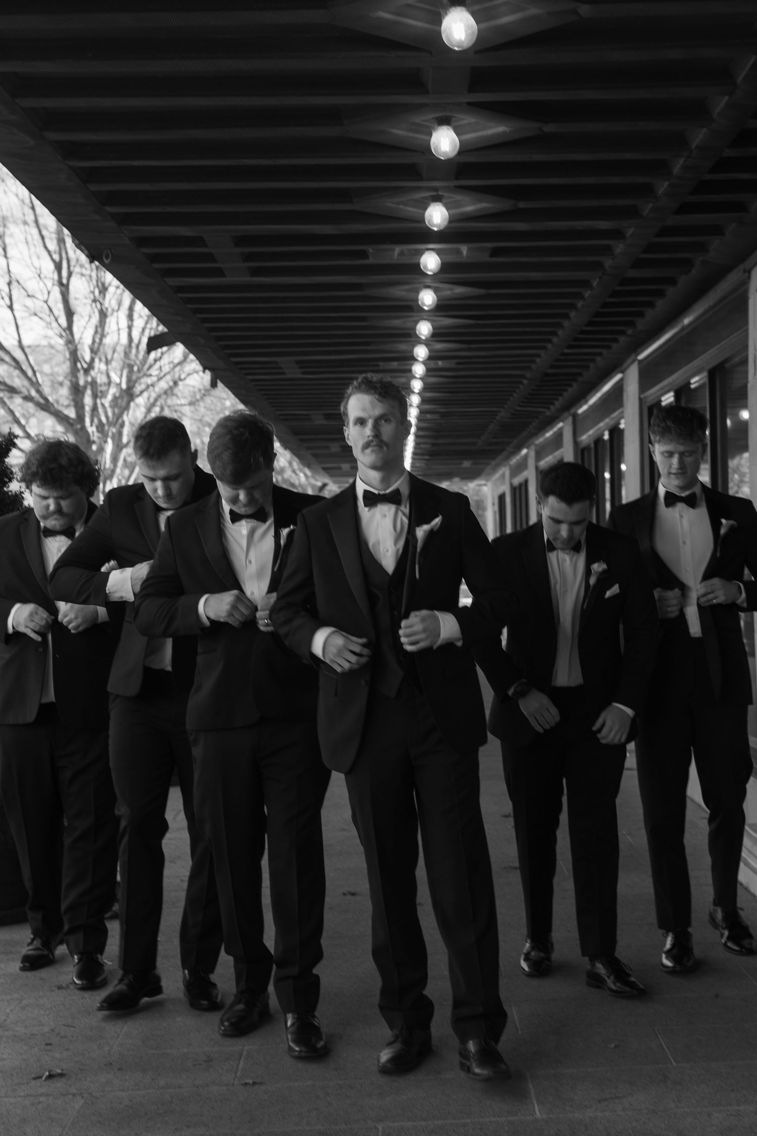 Groomsmen in black tuxedos stand in formation under dark beams with overhead lighting.