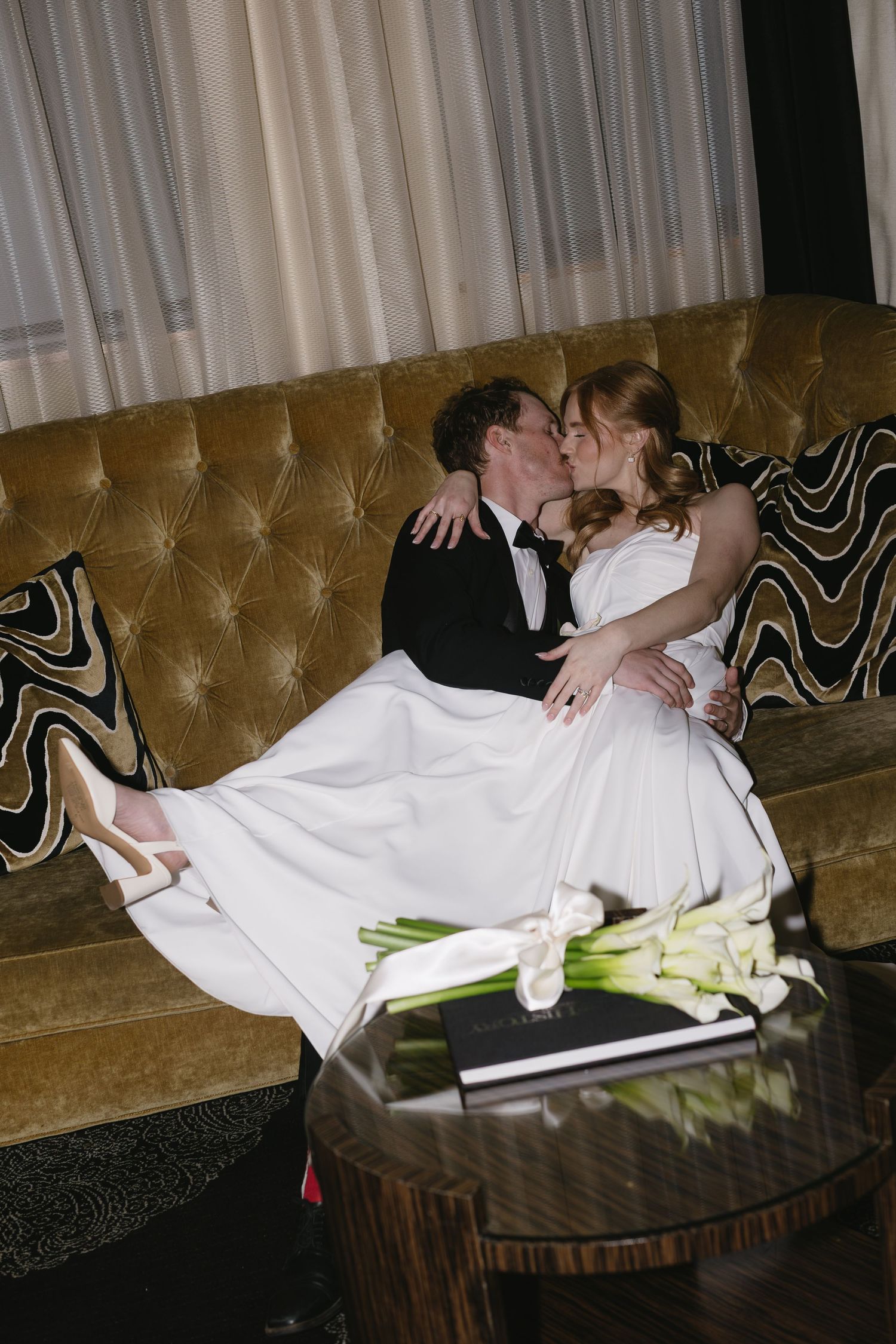 A couple relaxes on an elegant tufted couch with white calla lilies on the table.