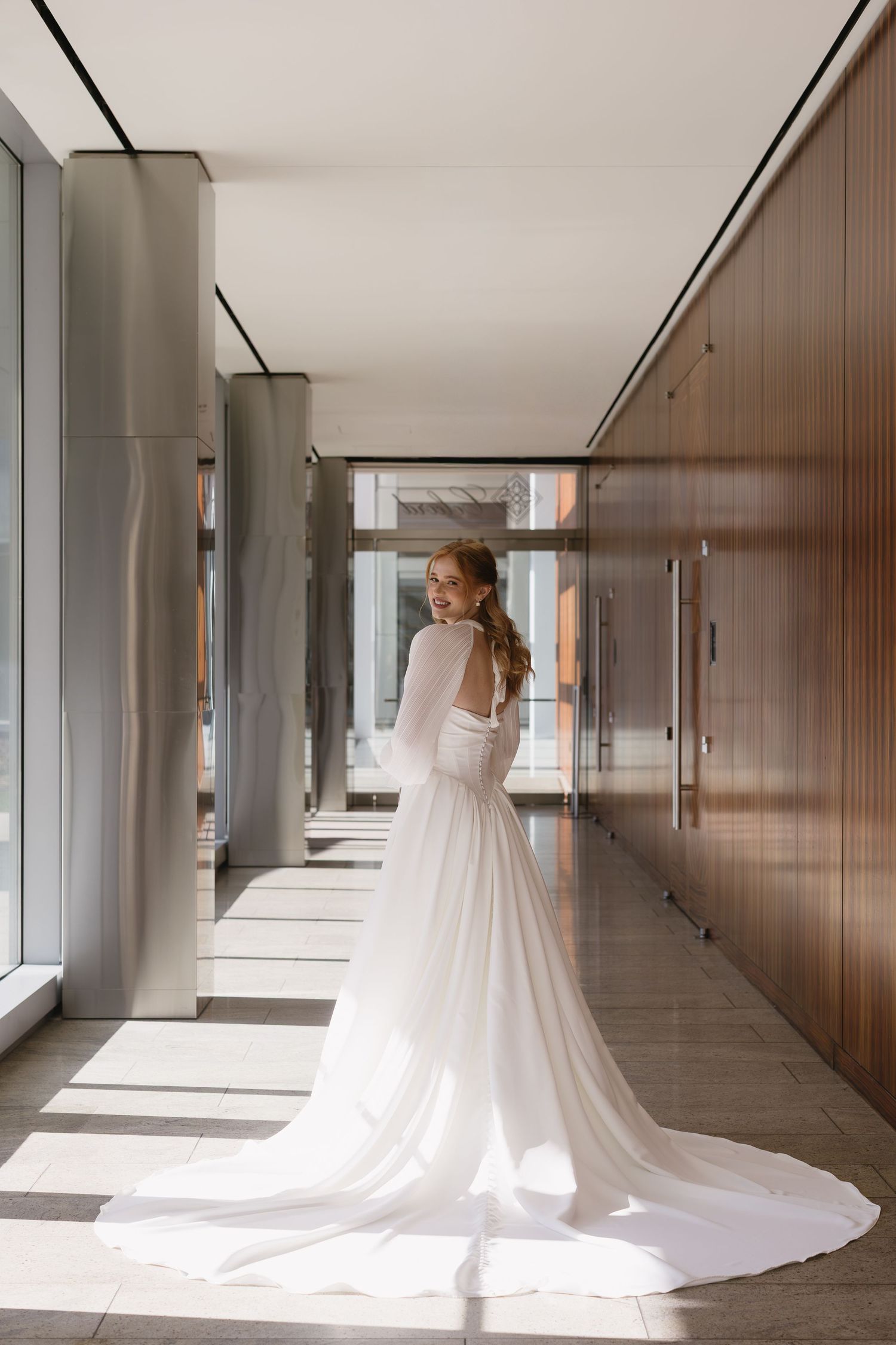 A flowing white wedding dress with long train photographed in a modern hallway with wood paneling.