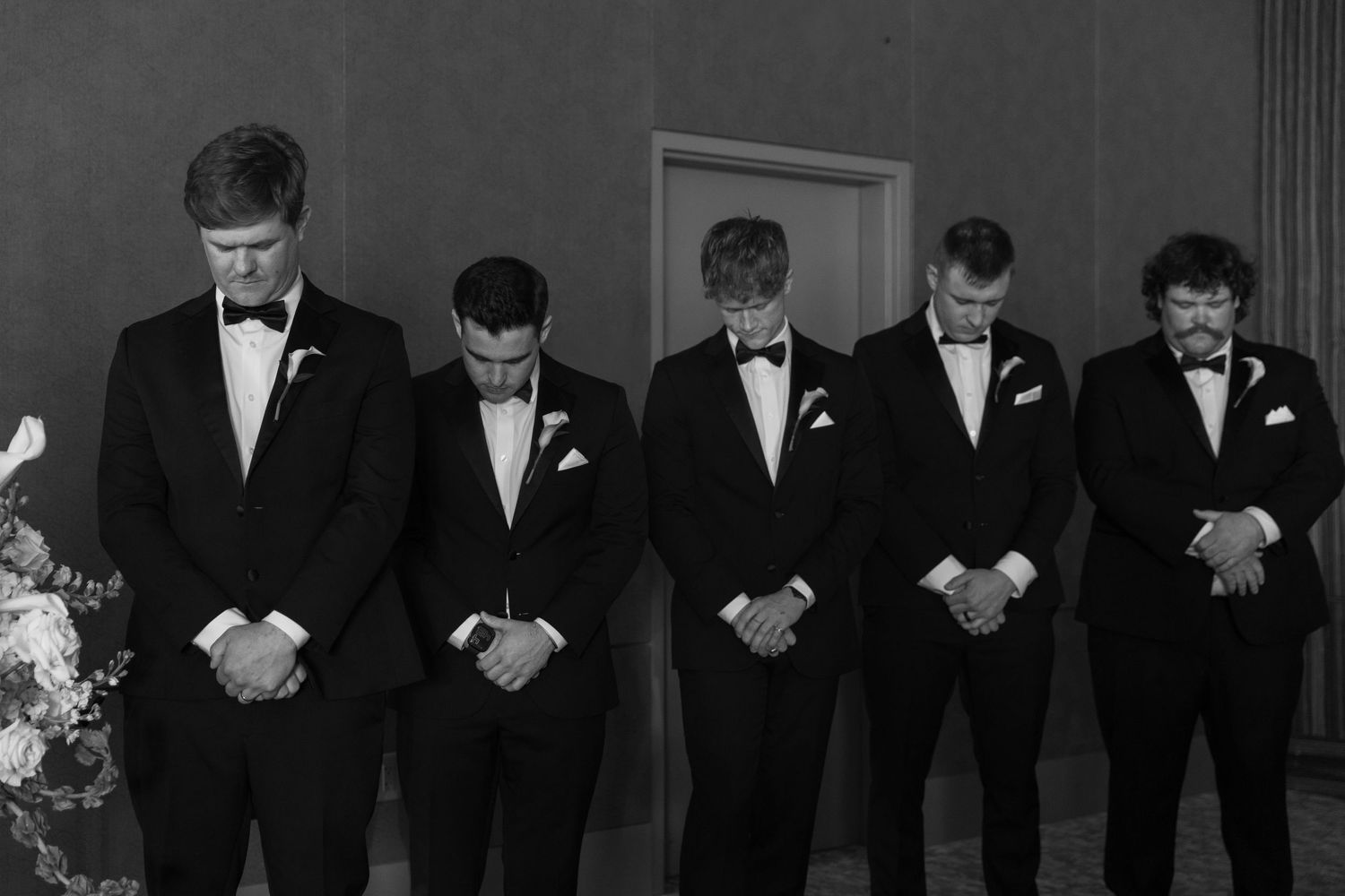 Groomsmen in black tuxedos stand in a line with heads bowed during a ceremony moment.