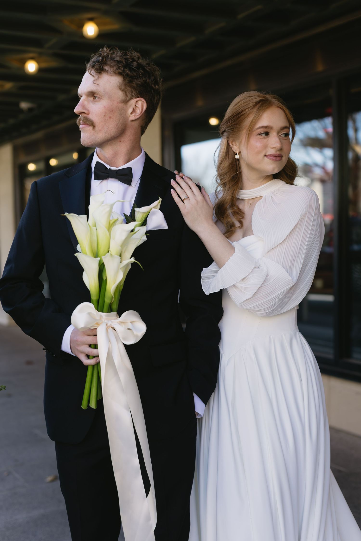 Wedding couple in classic attire with white calla lily bouquet against dark indoor backdrop.