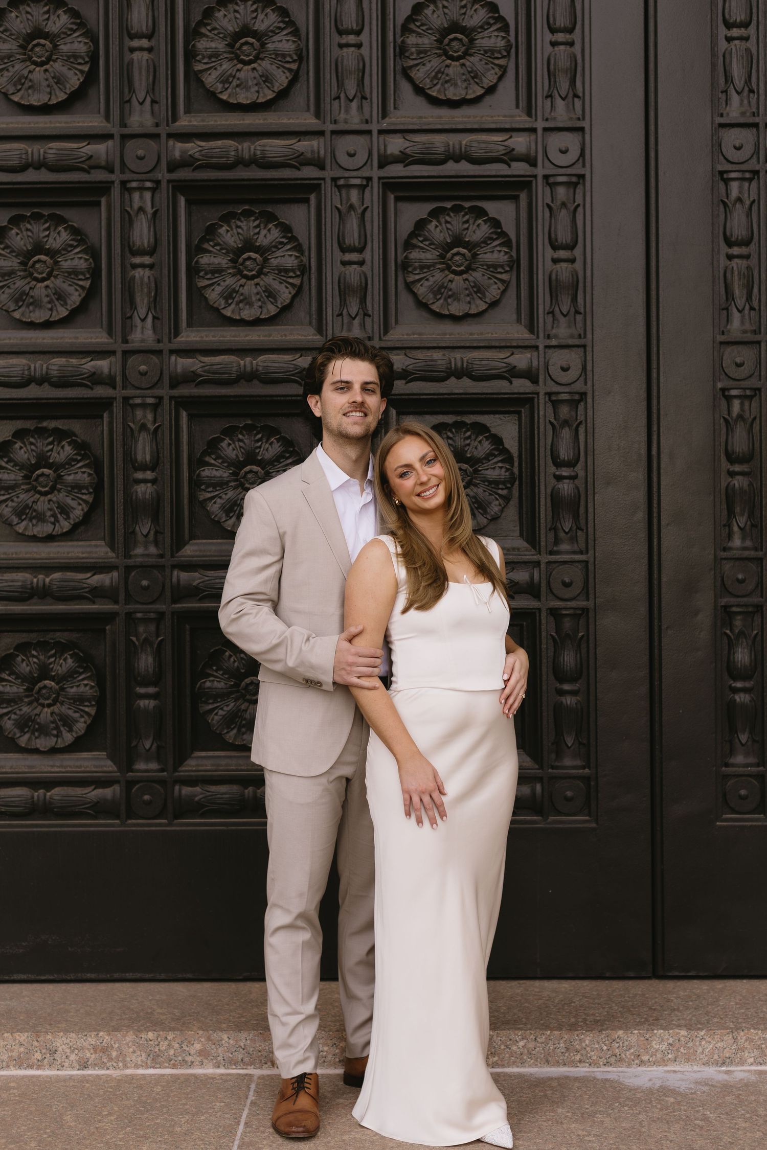 A couple in matching beige and white formal wear pose against an ornate dark door with floral patterns.
