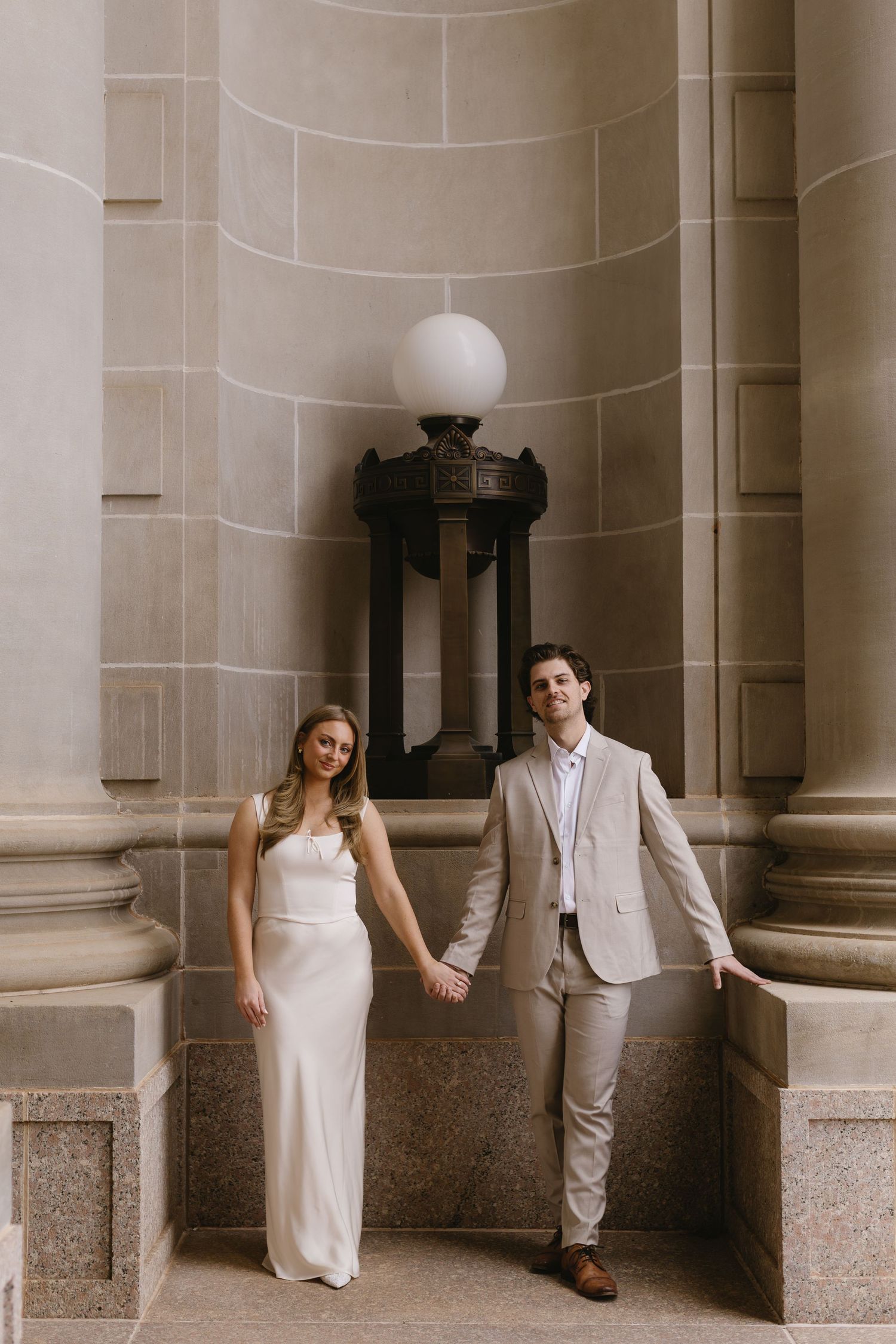 A couple in wedding attire pose between elegant stone columns with a vintage light fixture above.
