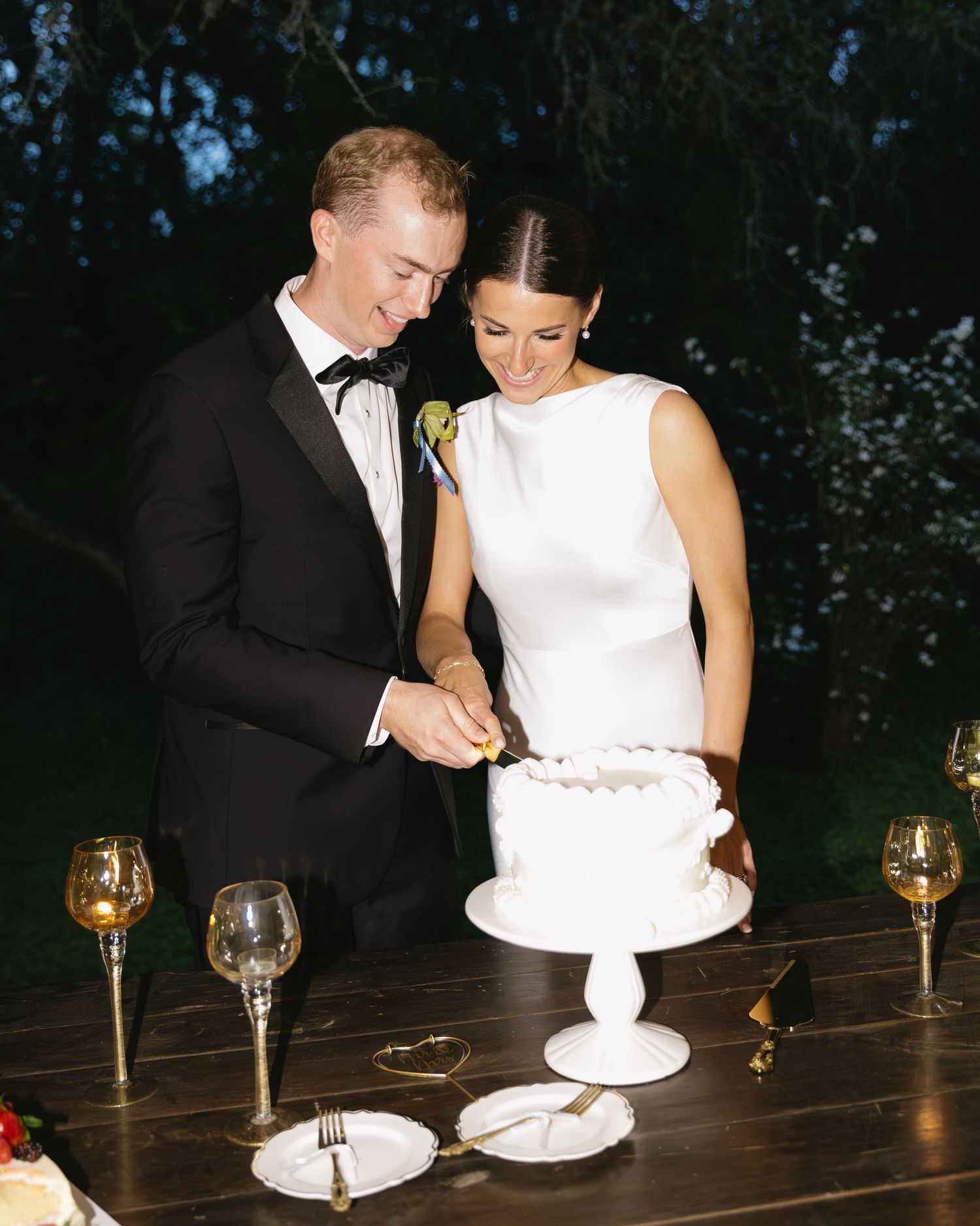 Newlyweds share cake cutting moment at reception with wine glasses on dark wooden table.