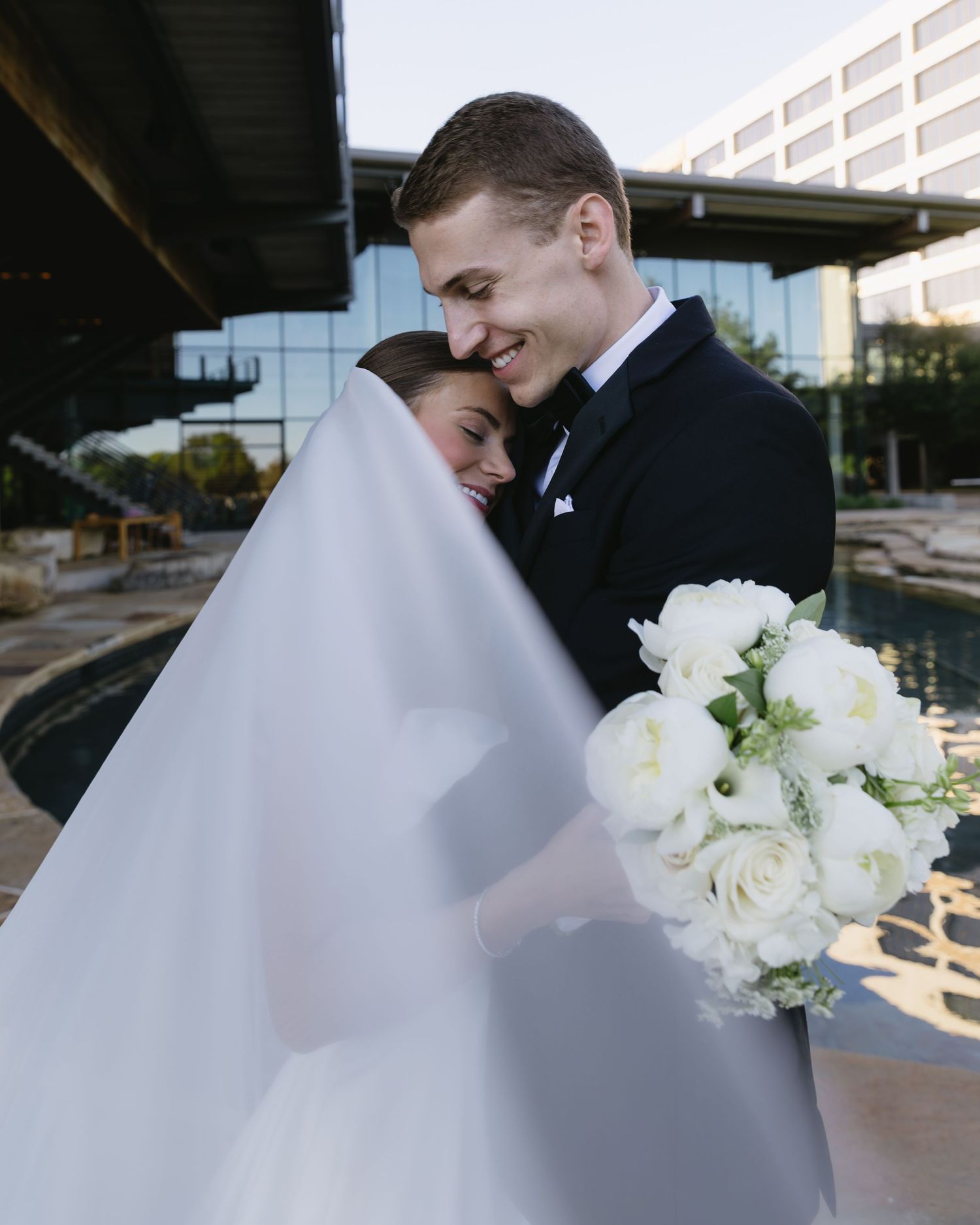 A romantic wedding couple embraces under dramatic lighting with the bride's veil flowing in the foreground.