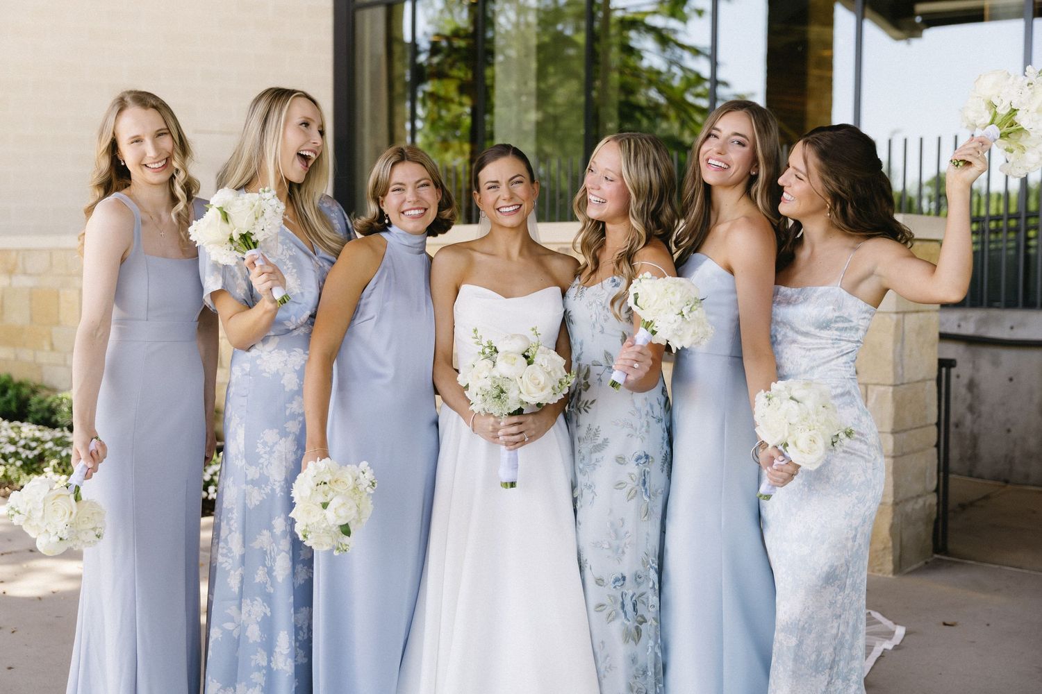 Bridesmaids in flowing light blue dresses pose together holding white bouquets.