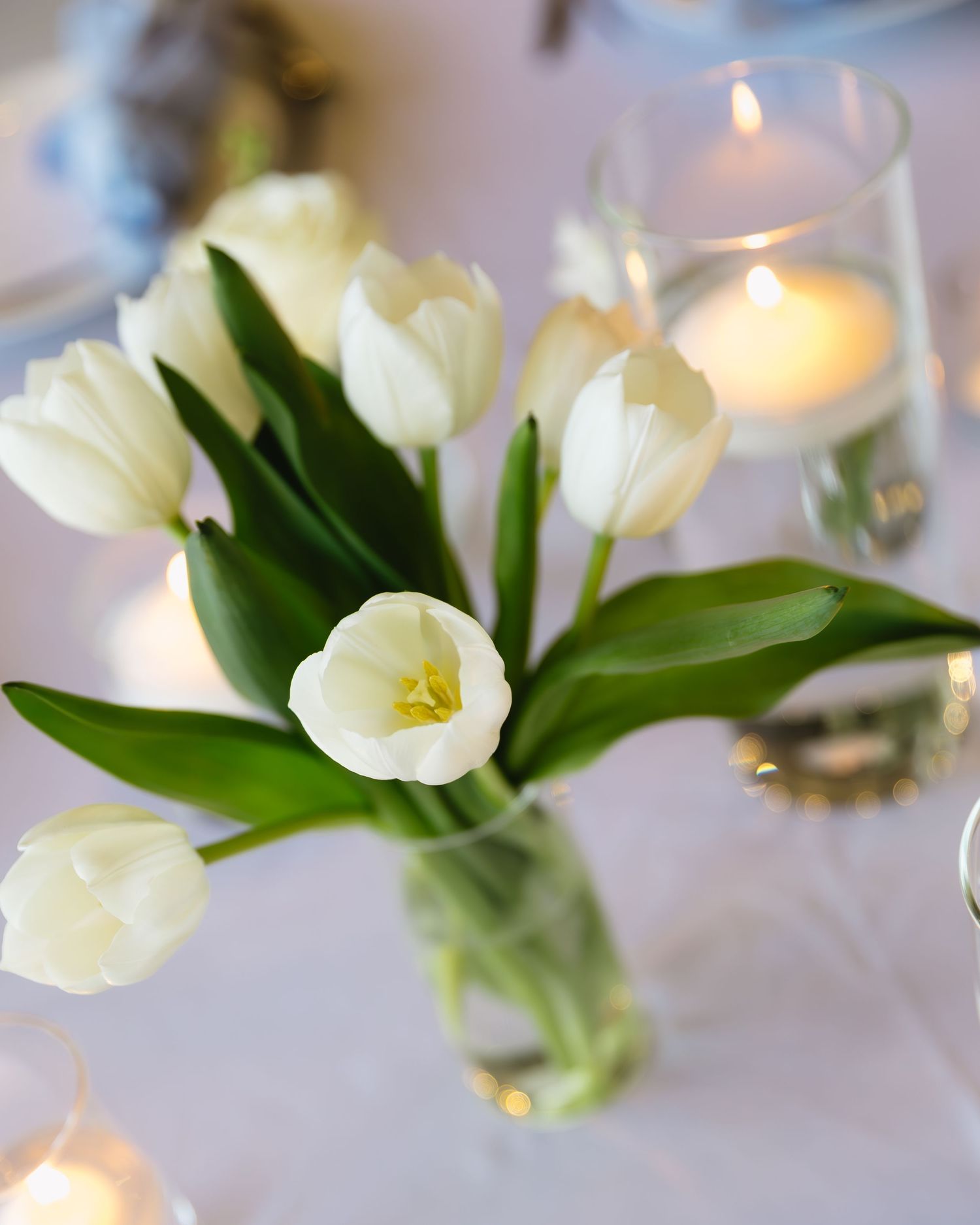 White tulips in a glass vase with glowing candlelight creating a romantic centerpiece setting.