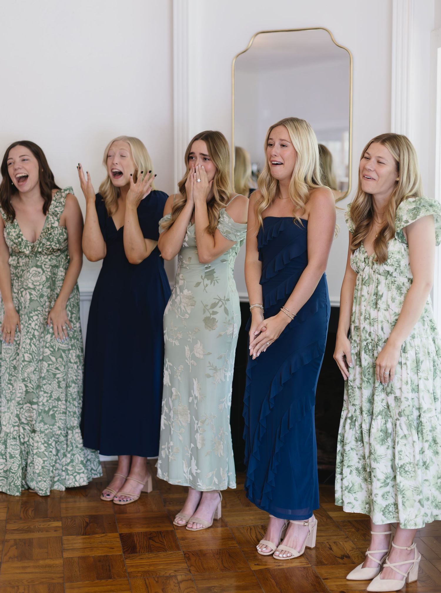 A group of women in floral and solid dresses laugh together in front of a mirror.