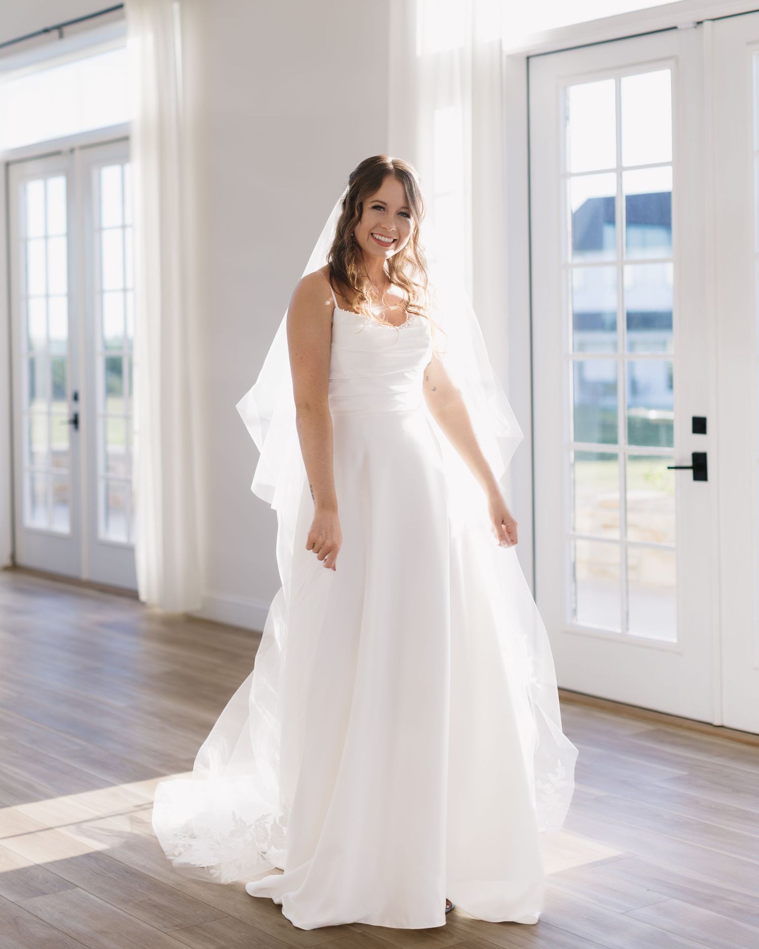 Bride in flowing white wedding dress twirling and dancing in a bright sunlit venue with hardwood floors.