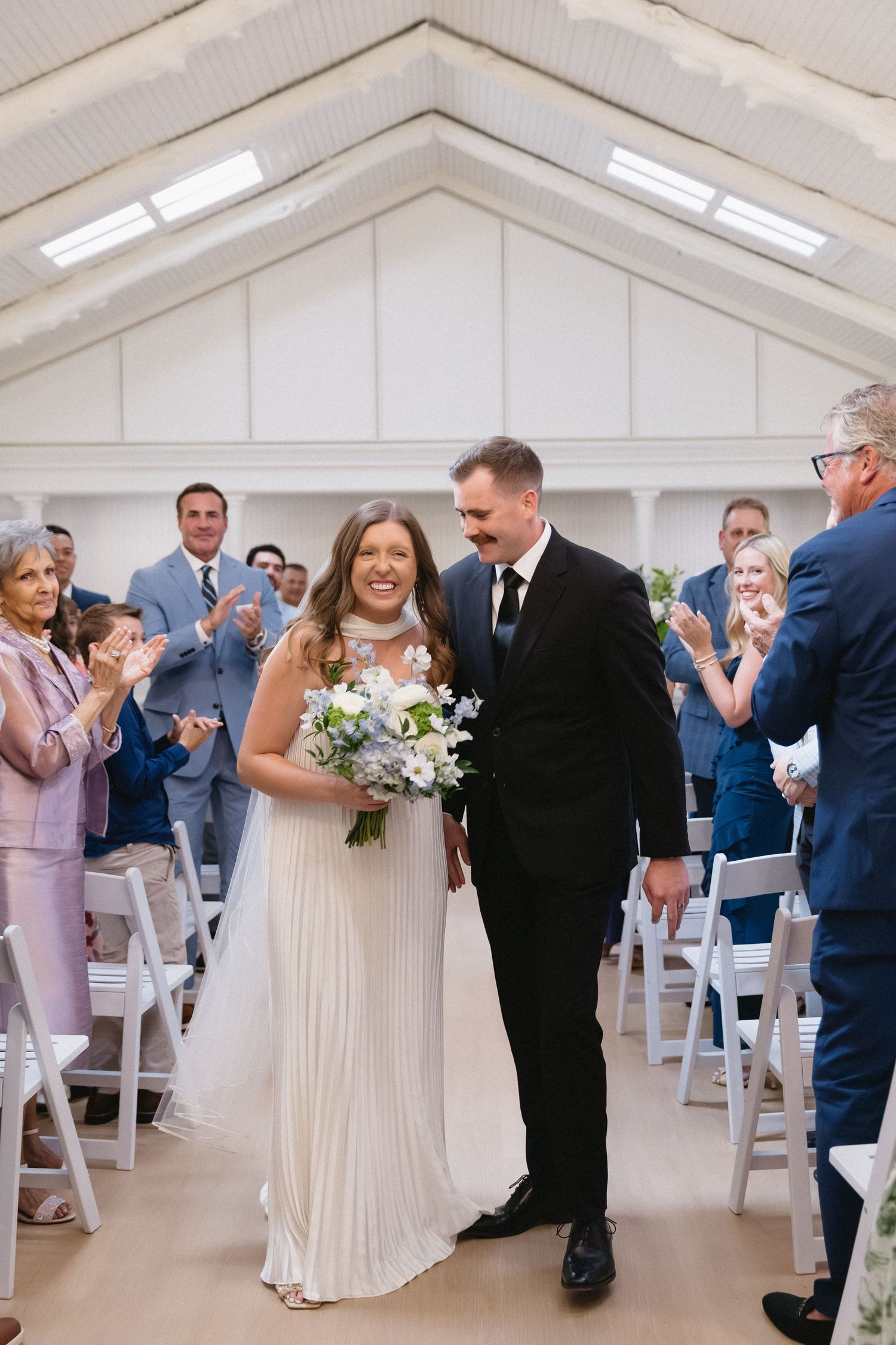 Wedding ceremony in a bright white chapel with guests seated in rows as couple exchanges vows.