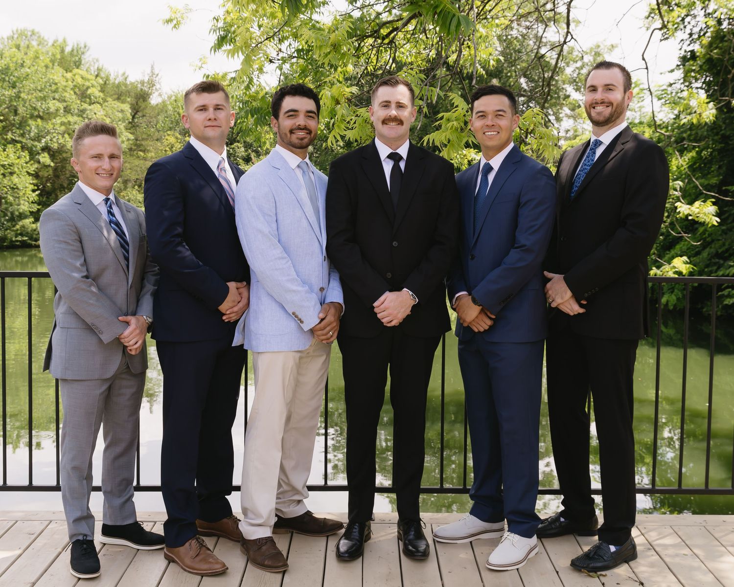 Group of groomsmen lined up on outdoor deck wearing various formal attire.