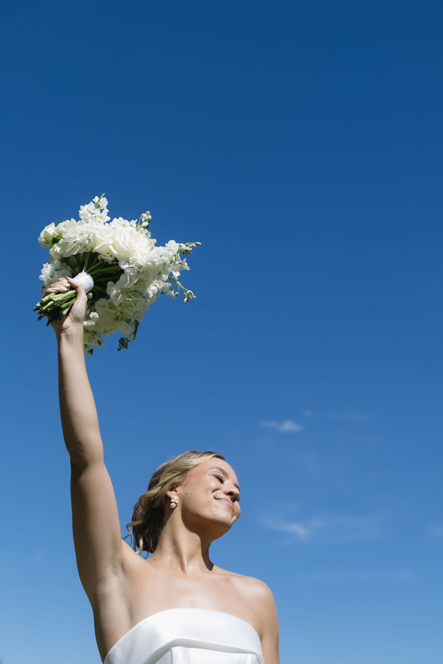 A person in a white dress holds up a white floral bouquet against a bright blue sky in celebration.