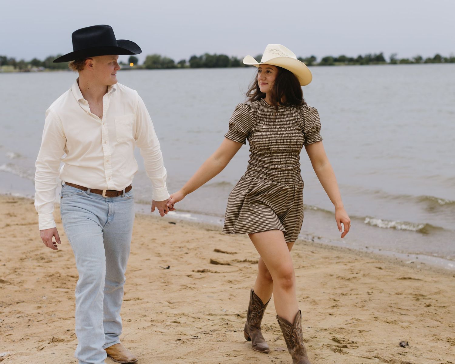 A couple playfully dances on a sandy beach wearing casual summer outfits.