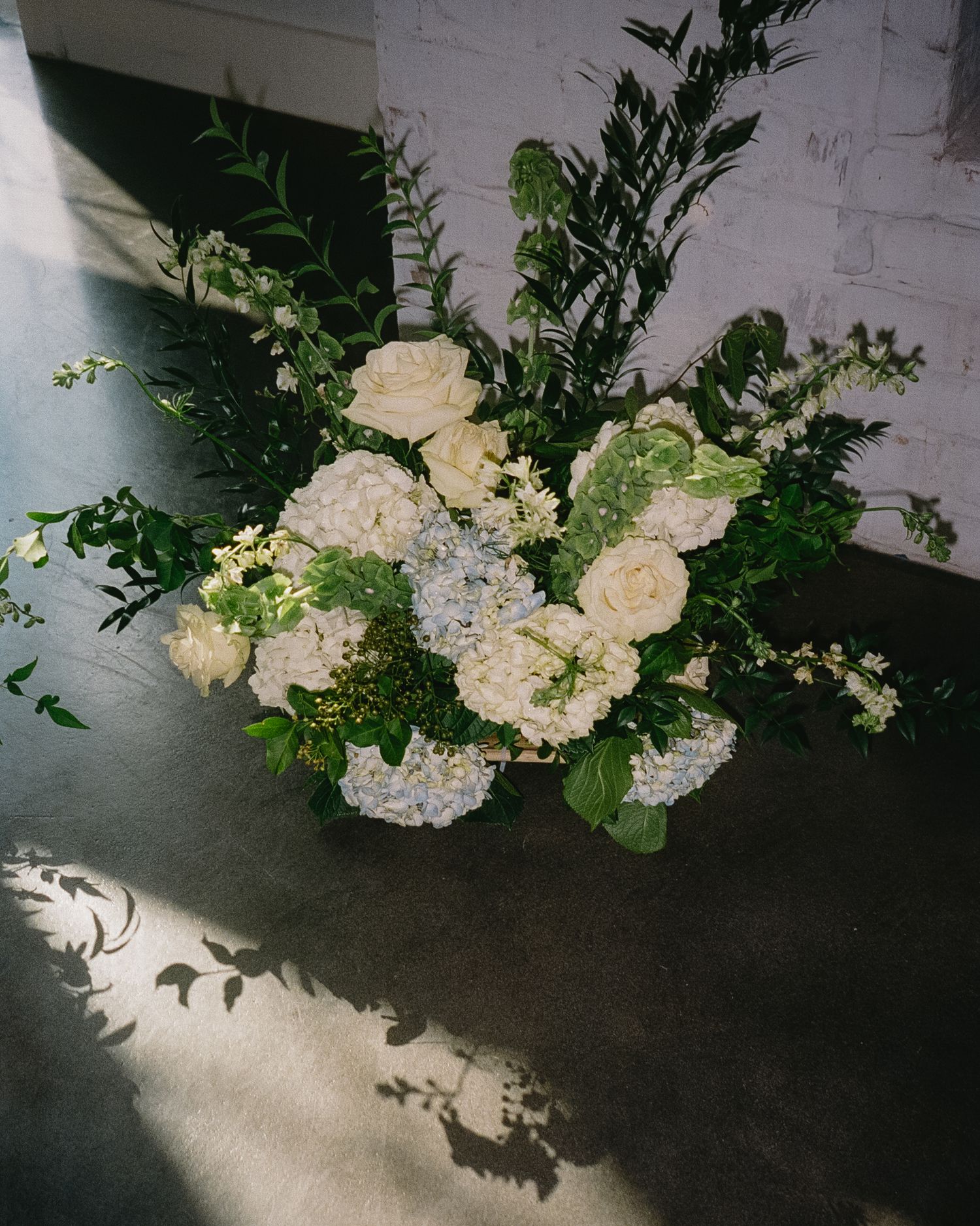 Elegant white and cream floral arrangement with greenery against white brick wall.