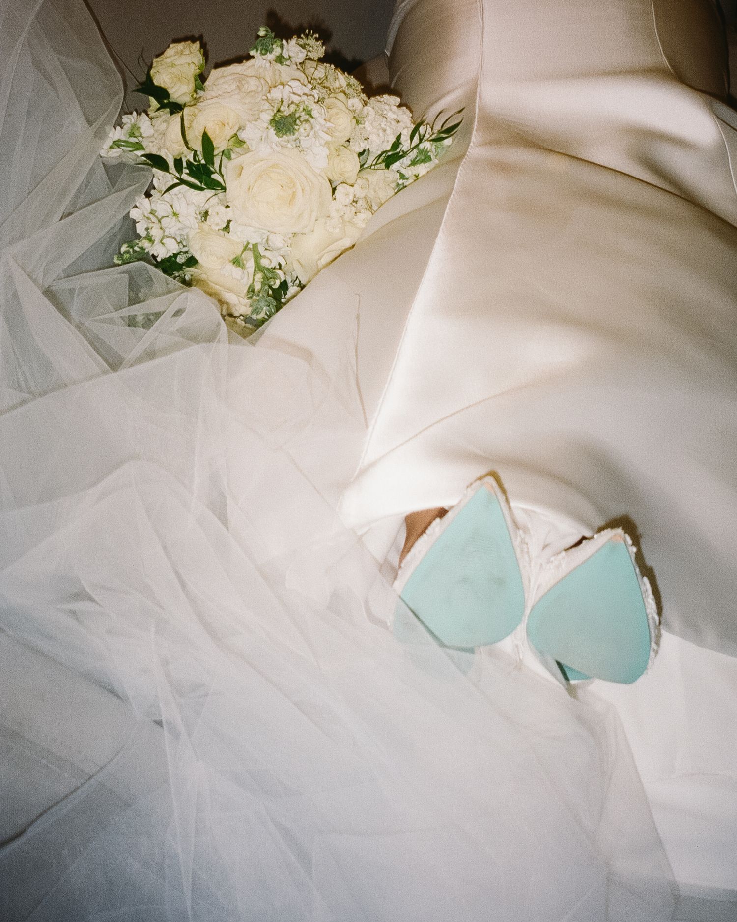 Wedding accessories including a white bouquet, ethereal veil fabric, and delicate light blue butterfly decorations rest on silk fabric.
