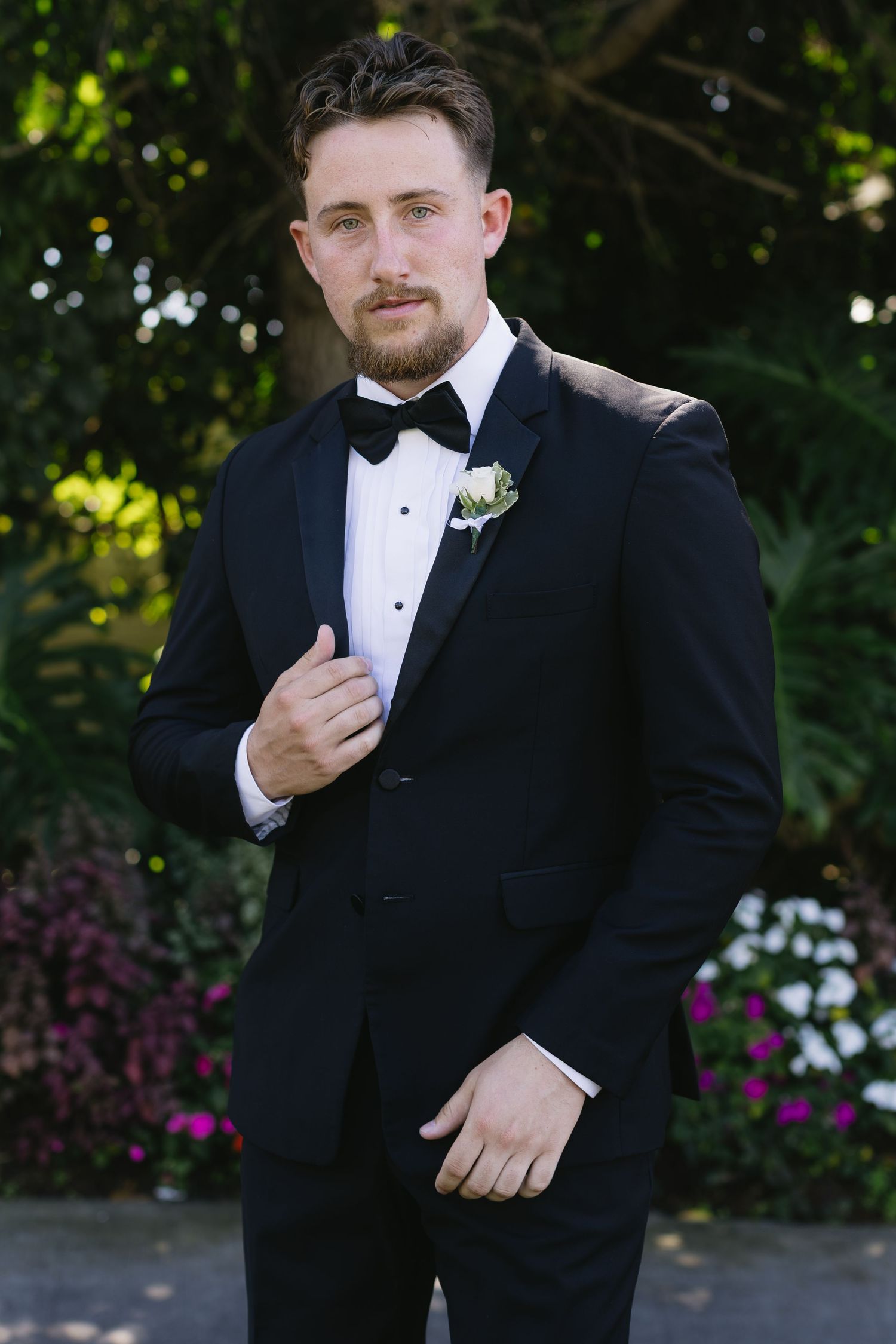Groom in classic black tuxedo and bowtie adjusting his collar outdoors.