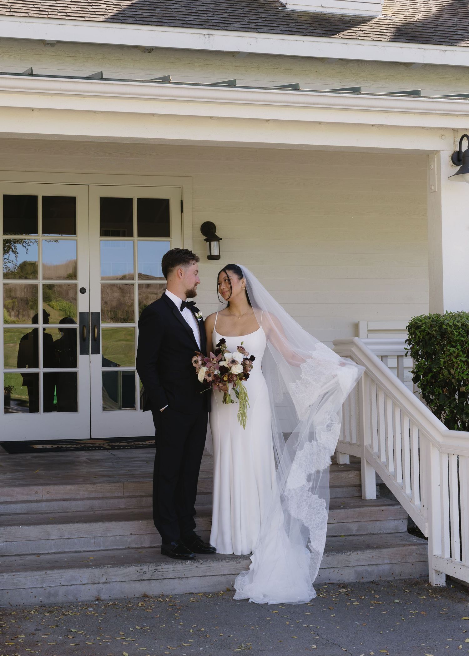 Wedding couple stands on white porch steps of elegant venue.