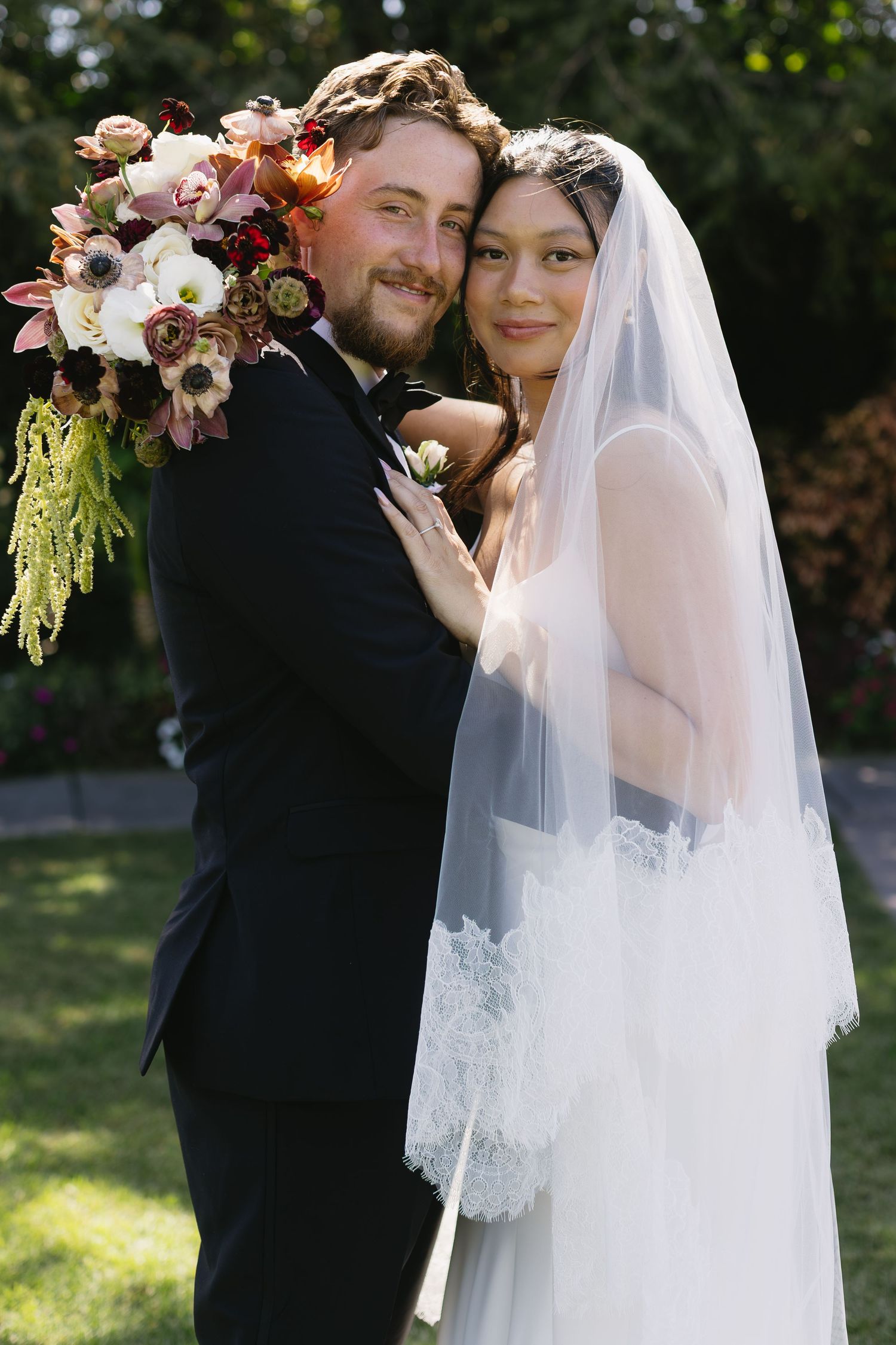 A romantic wedding couple poses outdoors with delicate pink floral arrangements and a flowing white veil.