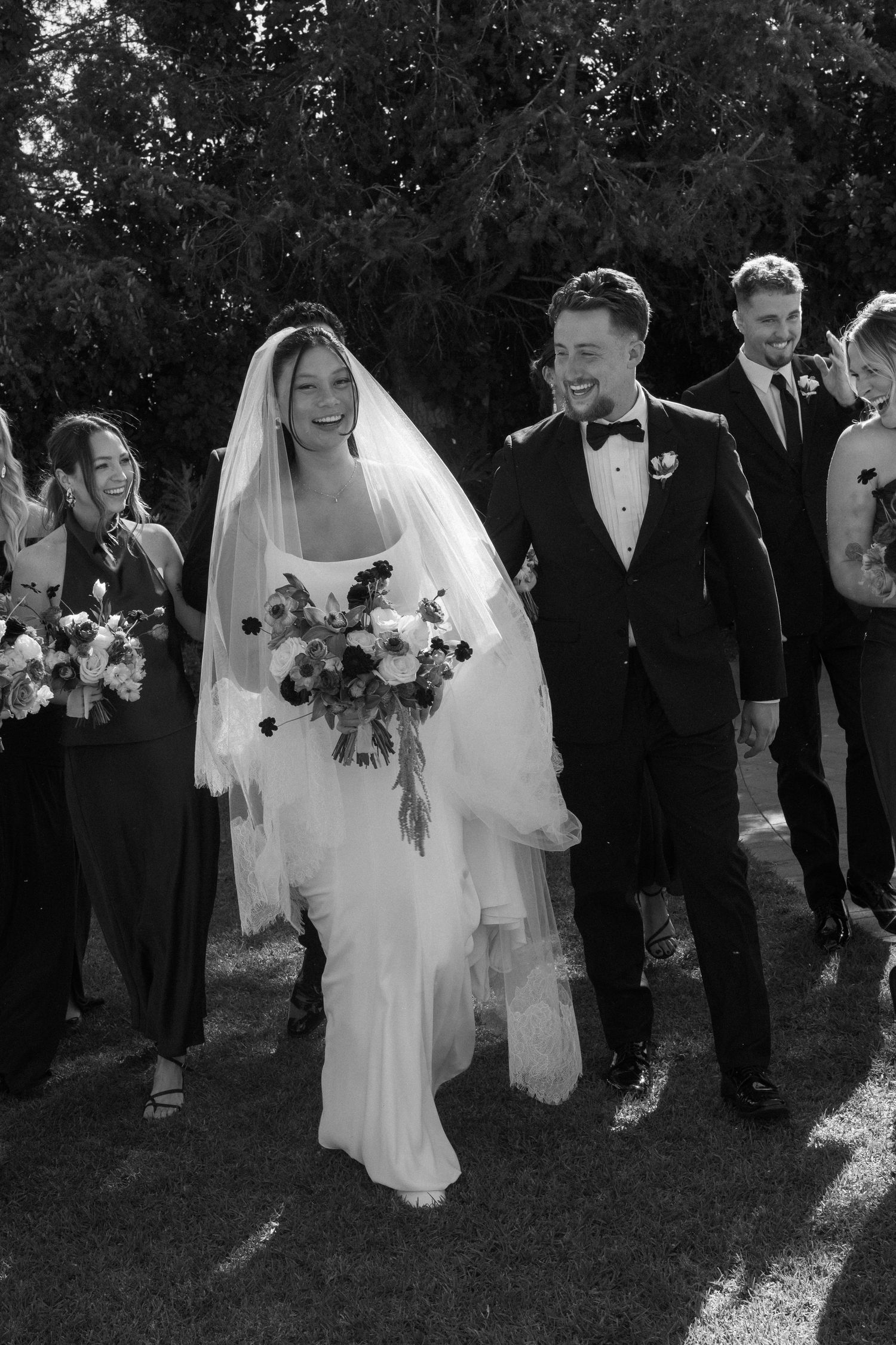 Wedding party walks together on grass with bride in white dress and attendants in formal attire.