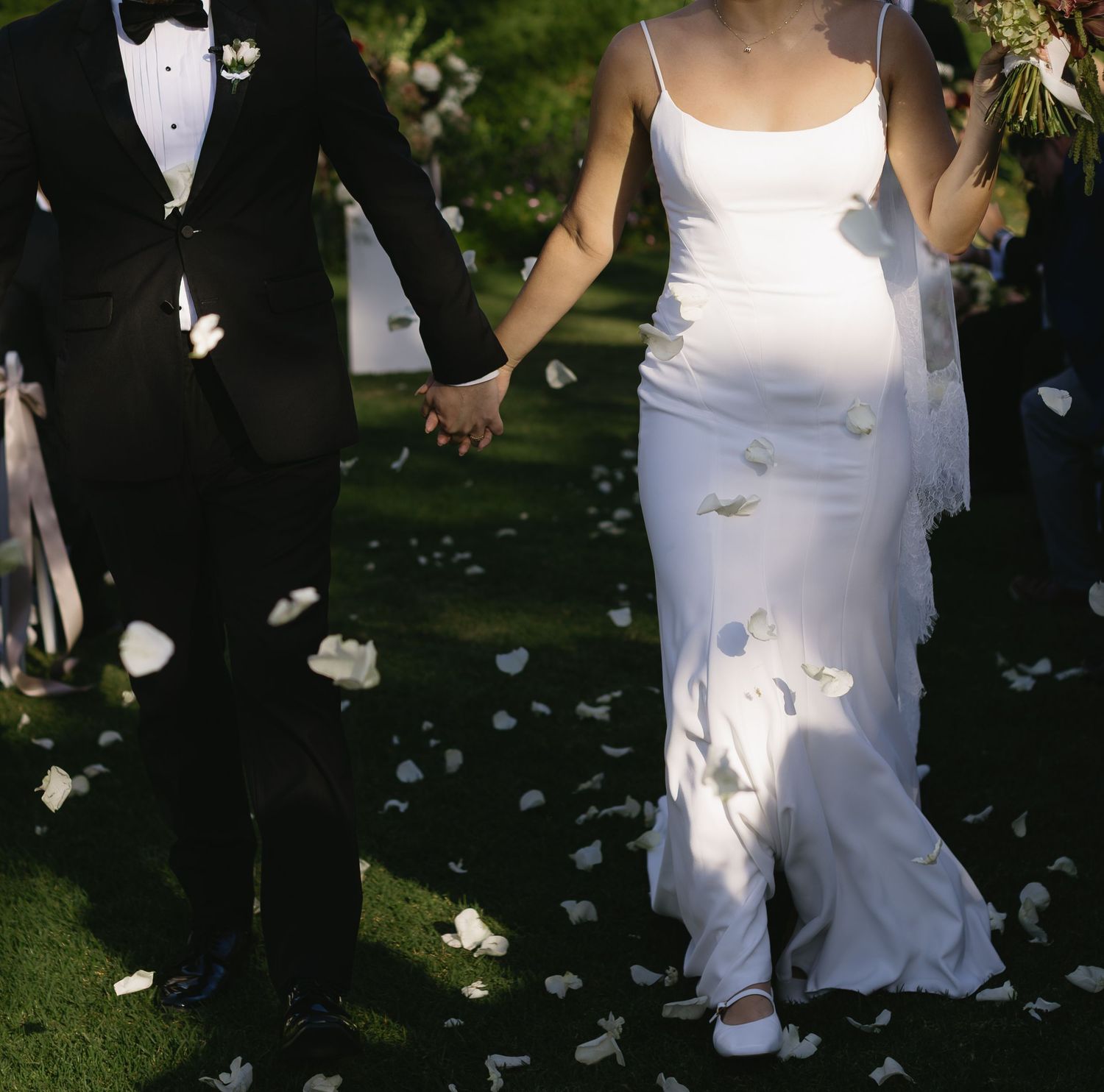 Wedding party members in formal attire stand together outdoors while flower petals fall around them.