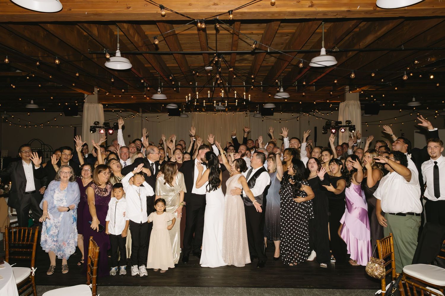 Wedding reception crowd celebrates with raised arms in an industrial-style venue with string lights.
