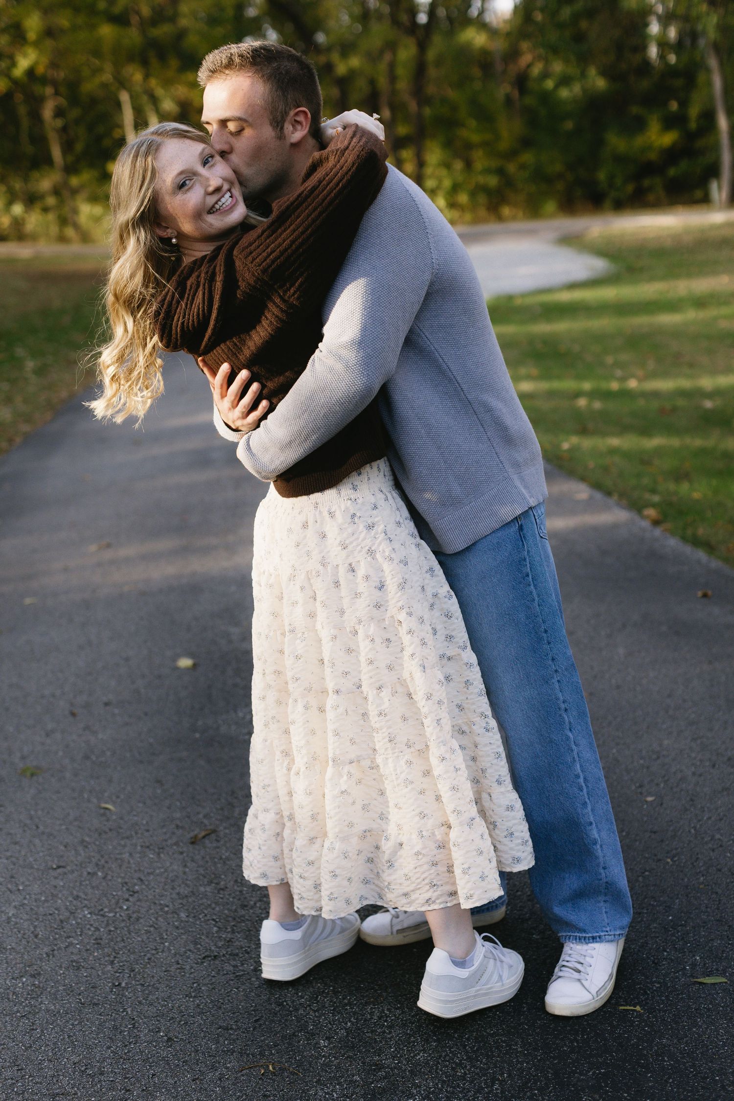 A romantic couple embraces on a paved path surrounded by autumn foliage.