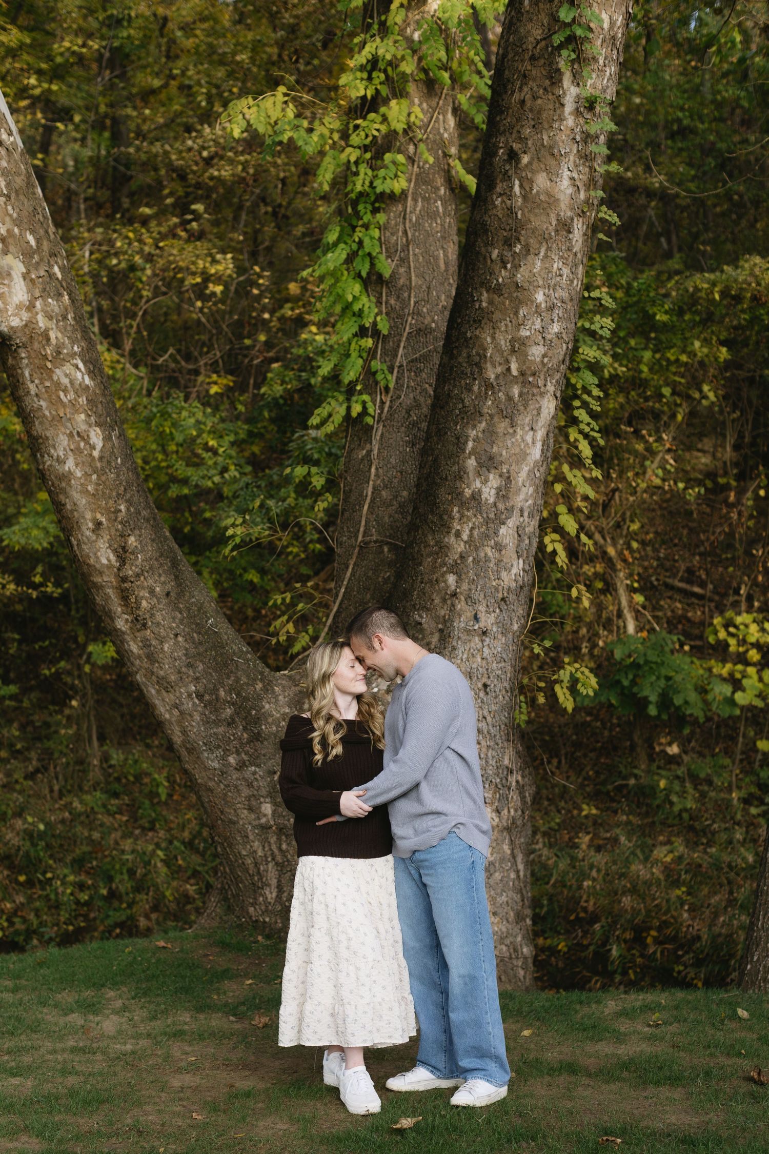 A couple embracing near large tree trunks in a wooded autumn setting.