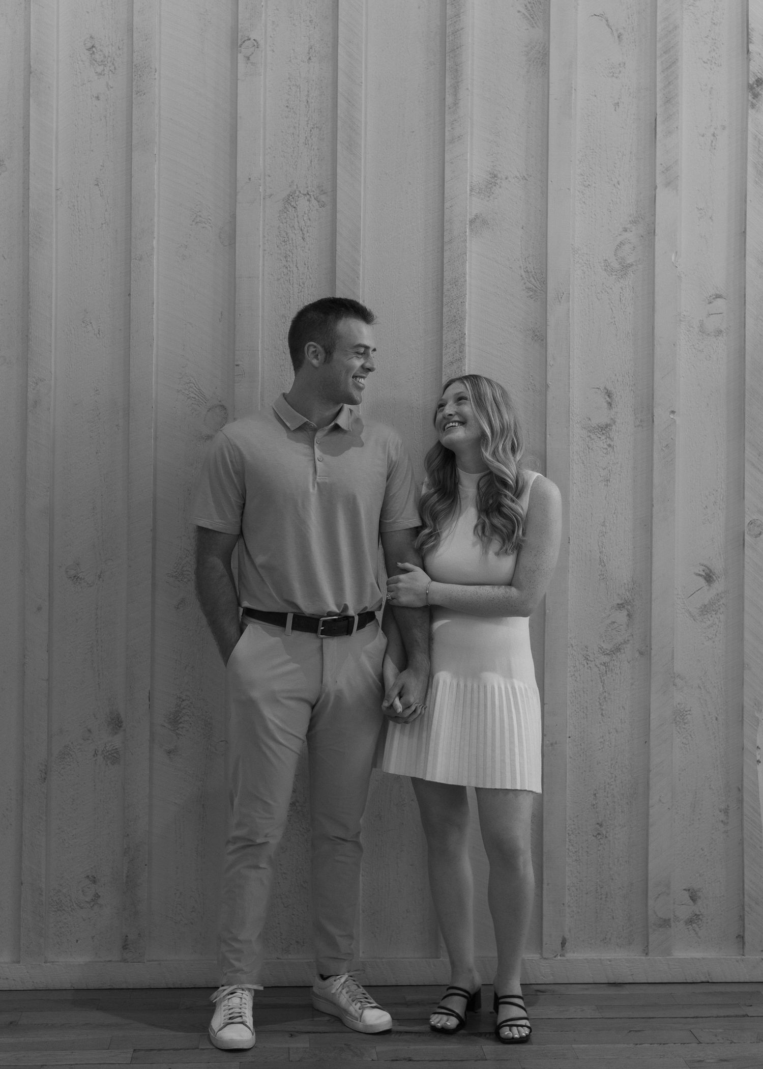A couple in casual attire poses against a corrugated metal wall in a black and white photograph.