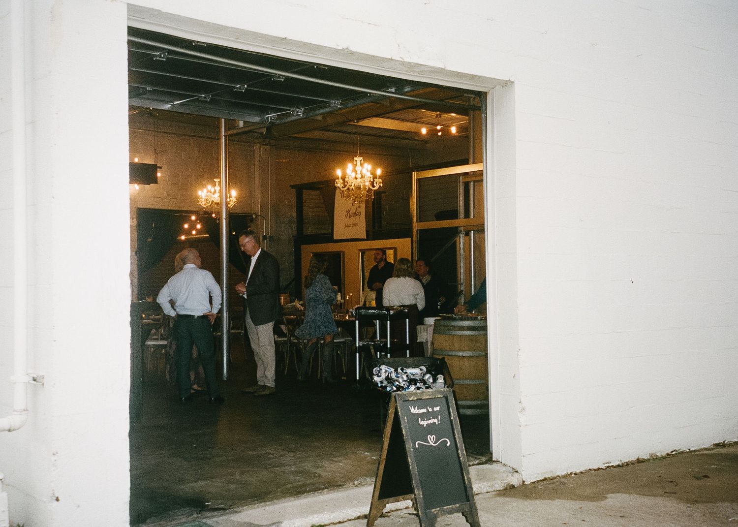 View through doorway of people gathering in dimly lit venue with chalkboard sign.