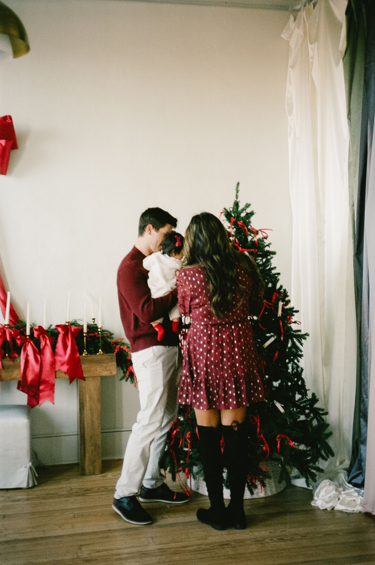 Holiday photo session with a decorated Christmas tree and red bows in a rustic indoor setting.