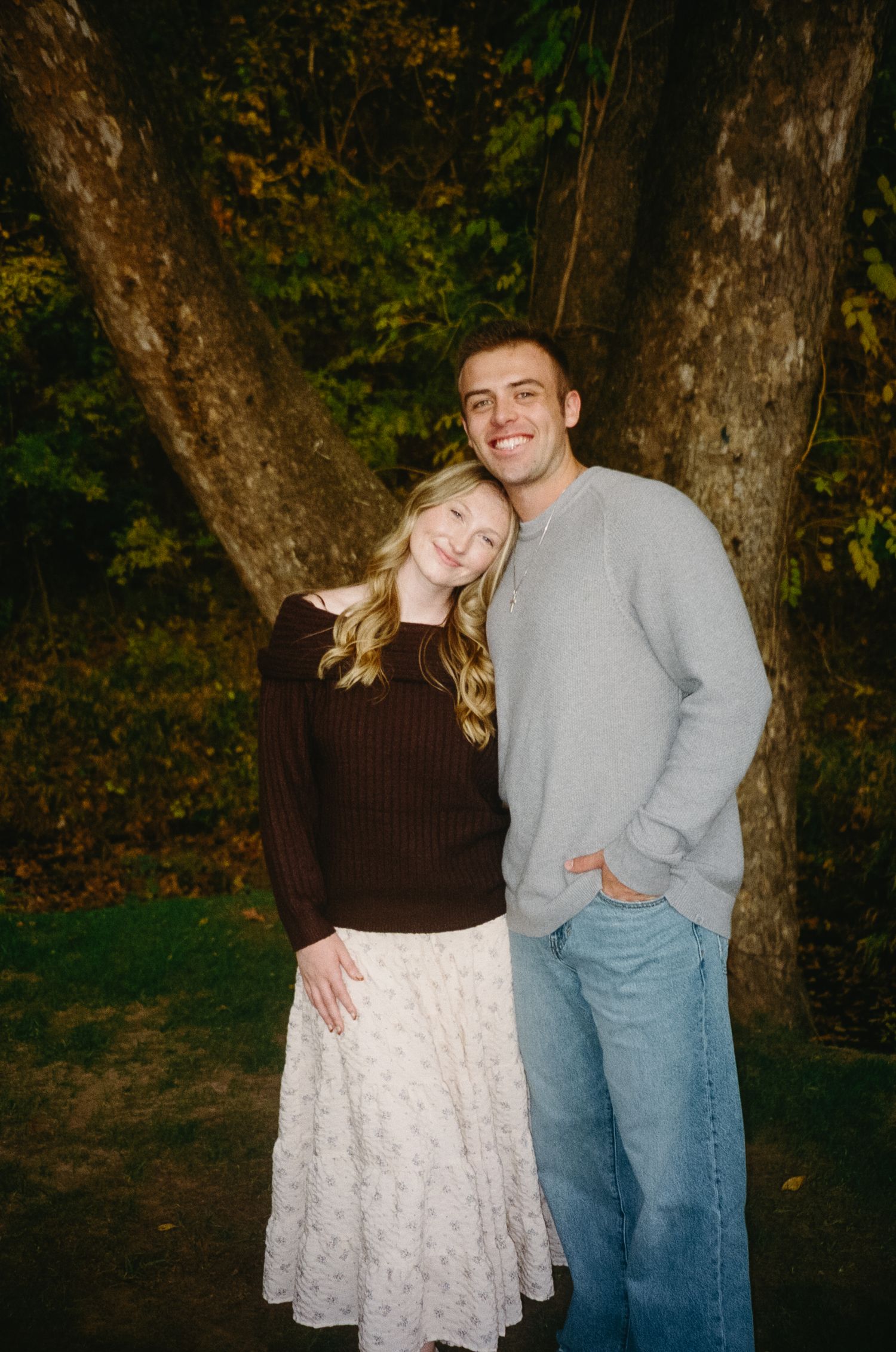 Couple in casual attire pose together against autumn trees in a park setting.