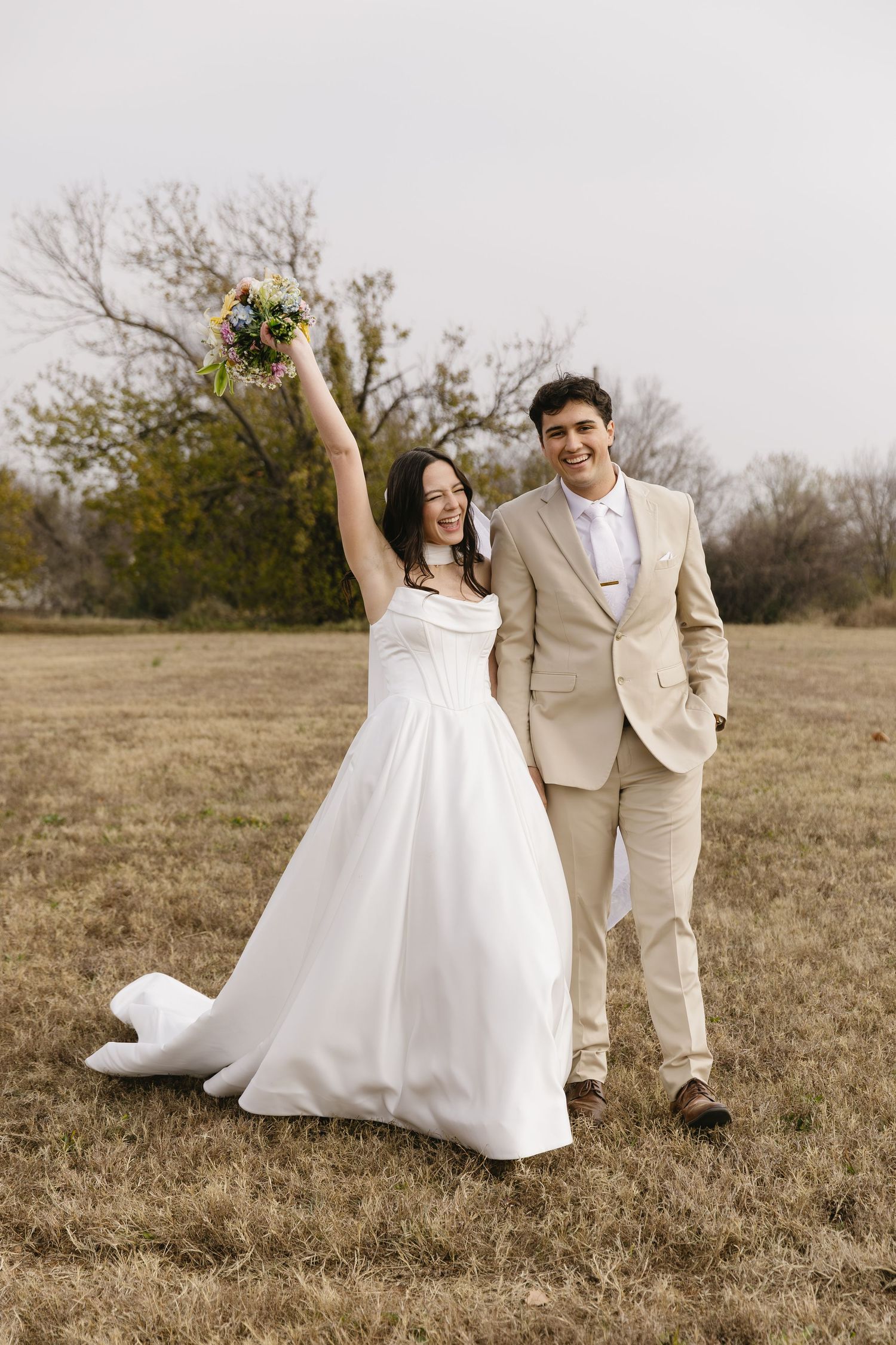 Playful wedding couple poses in a rustic field with the bride twirling her white dress and groom in beige suit.