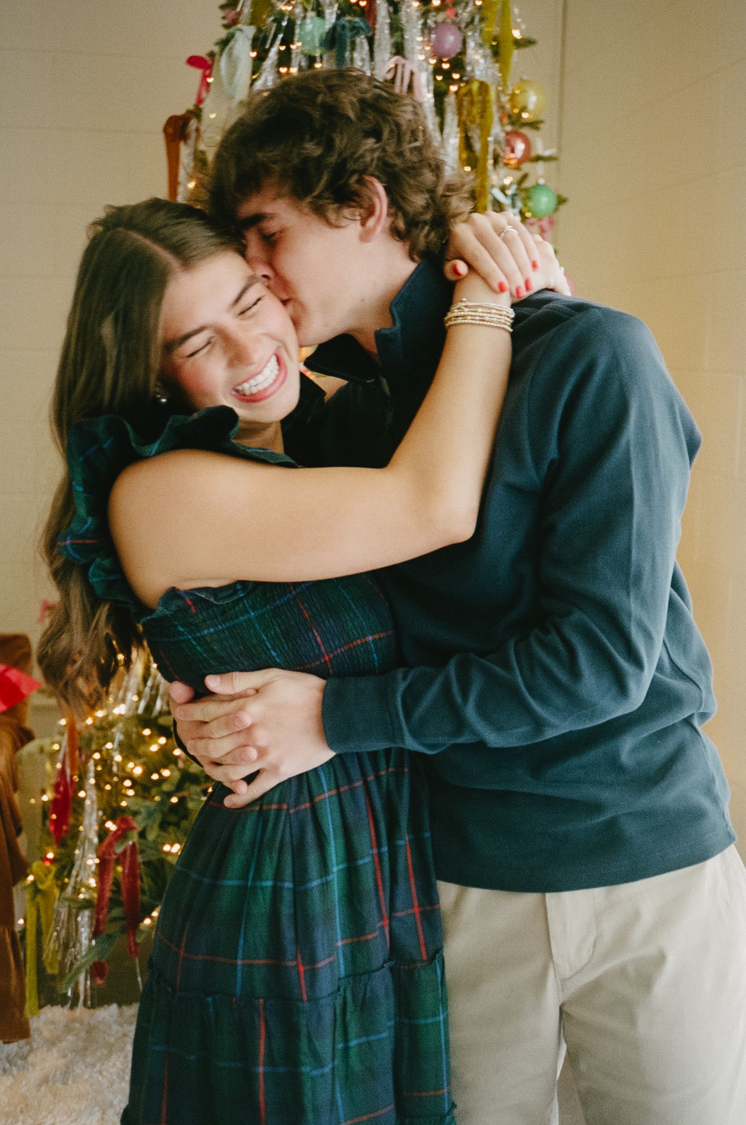 A romantic couple shares an intimate moment in front of a Christmas-decorated wall with twinkling lights.