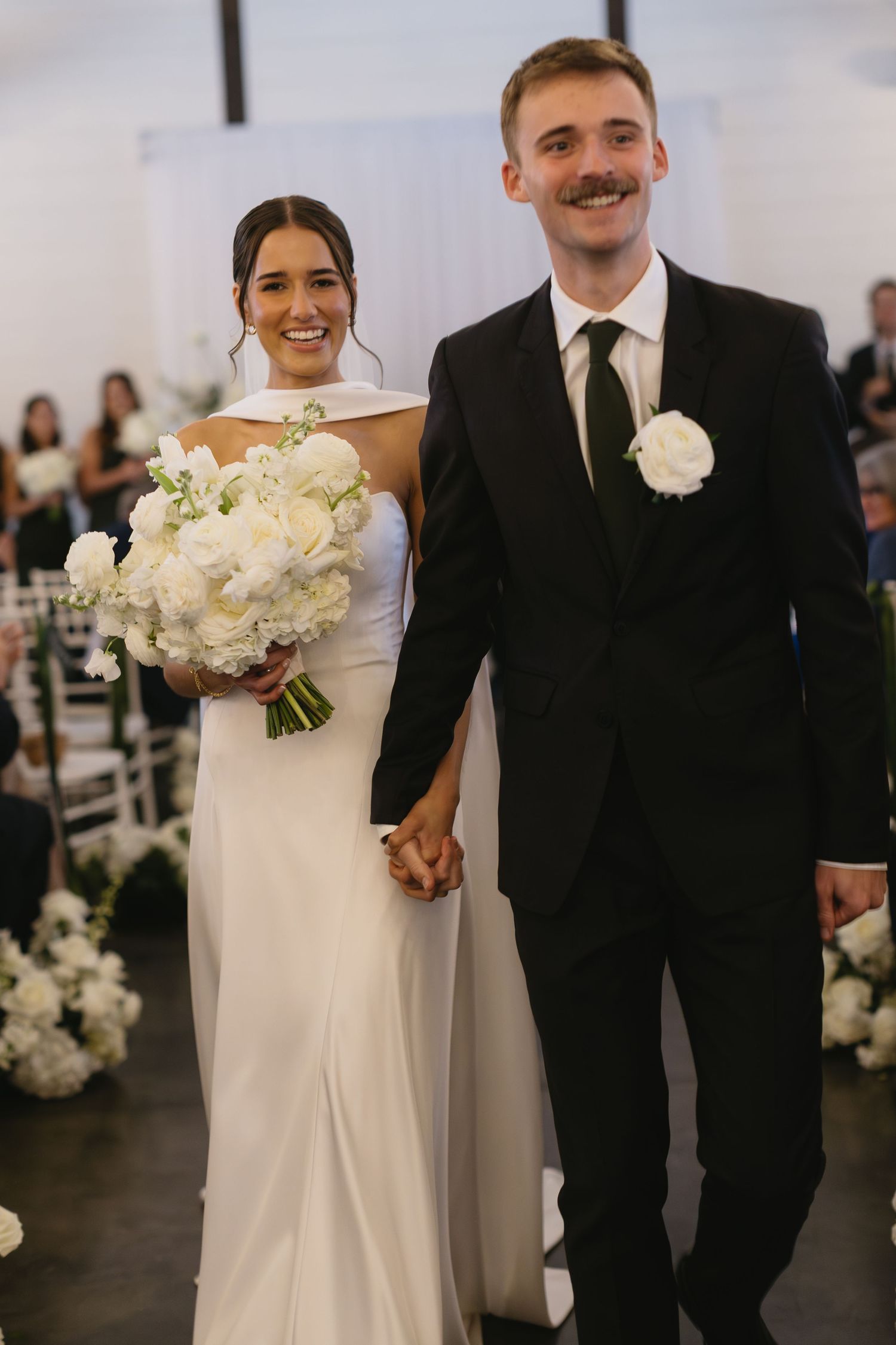 A wedding couple walks down the aisle holding hands with white floral decorations.