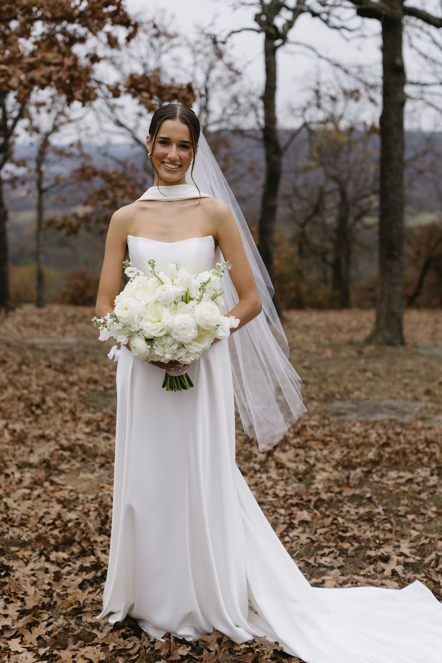 A bride holds a white bouquet while posing in an autumn forest setting.