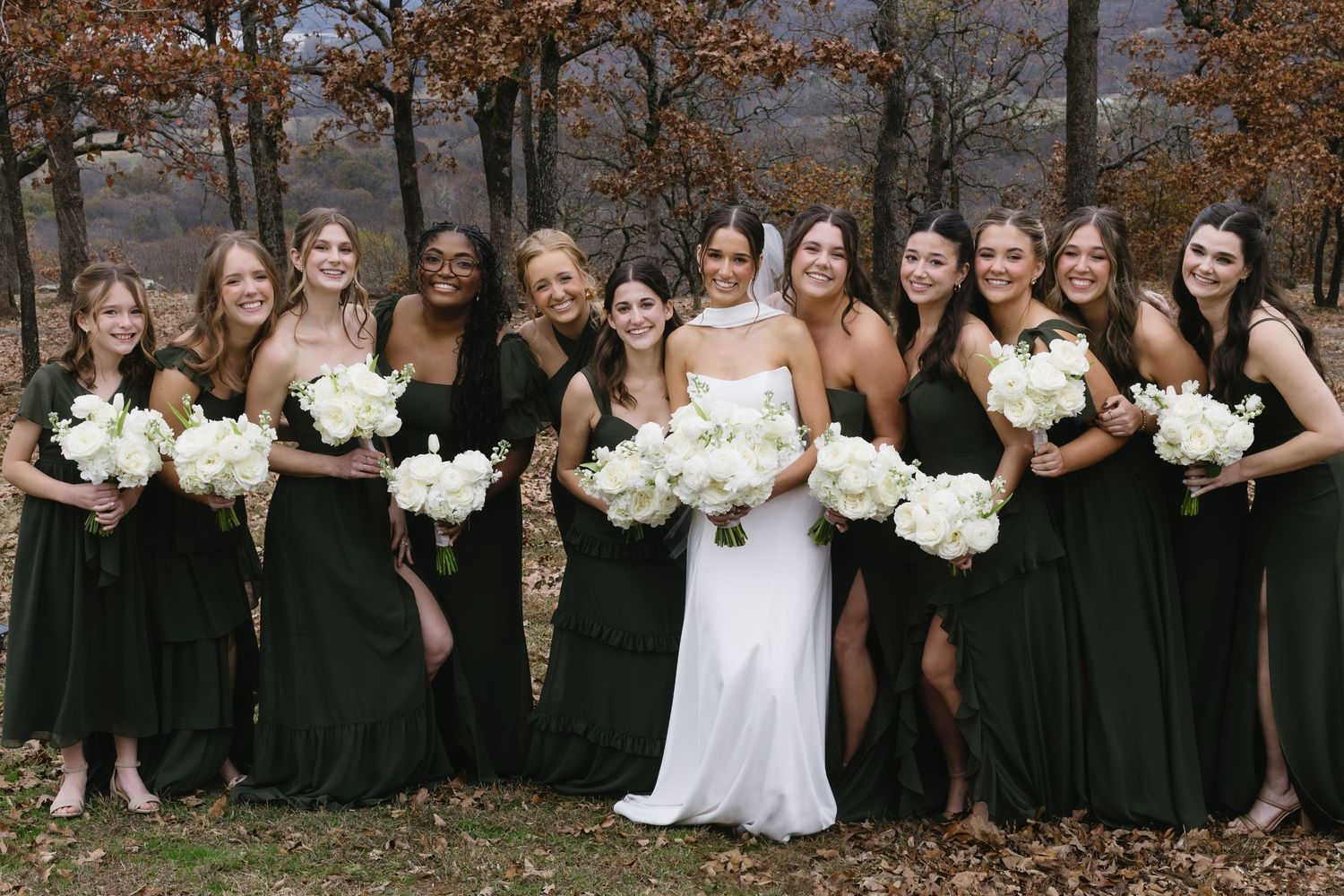 Wedding party in black dresses and white bouquets pose together in a fall outdoor setting.