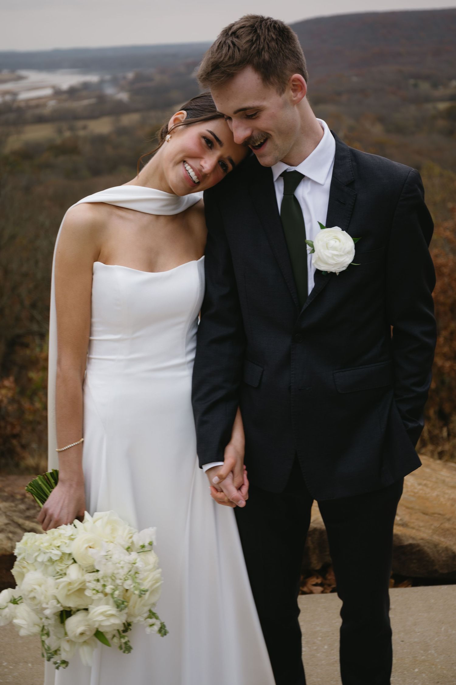 Elegant wedding couple shares intimate moments against a scenic hillside backdrop while holding white floral bouquet.