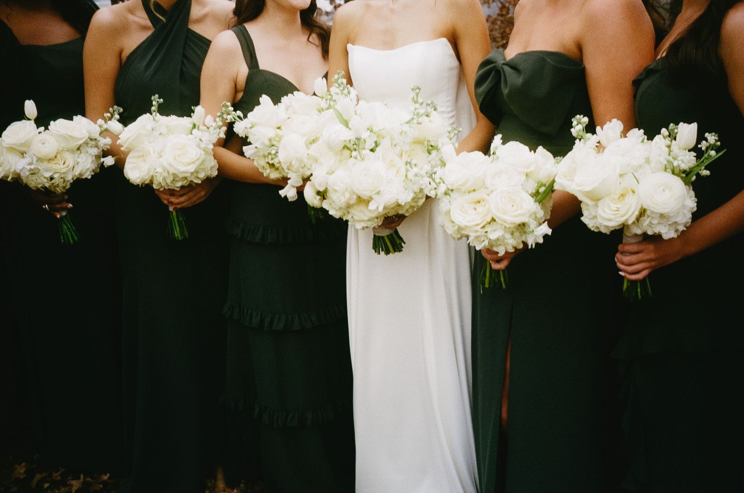 Bridesmaids in black dresses holding white floral bouquets next to bride in white.