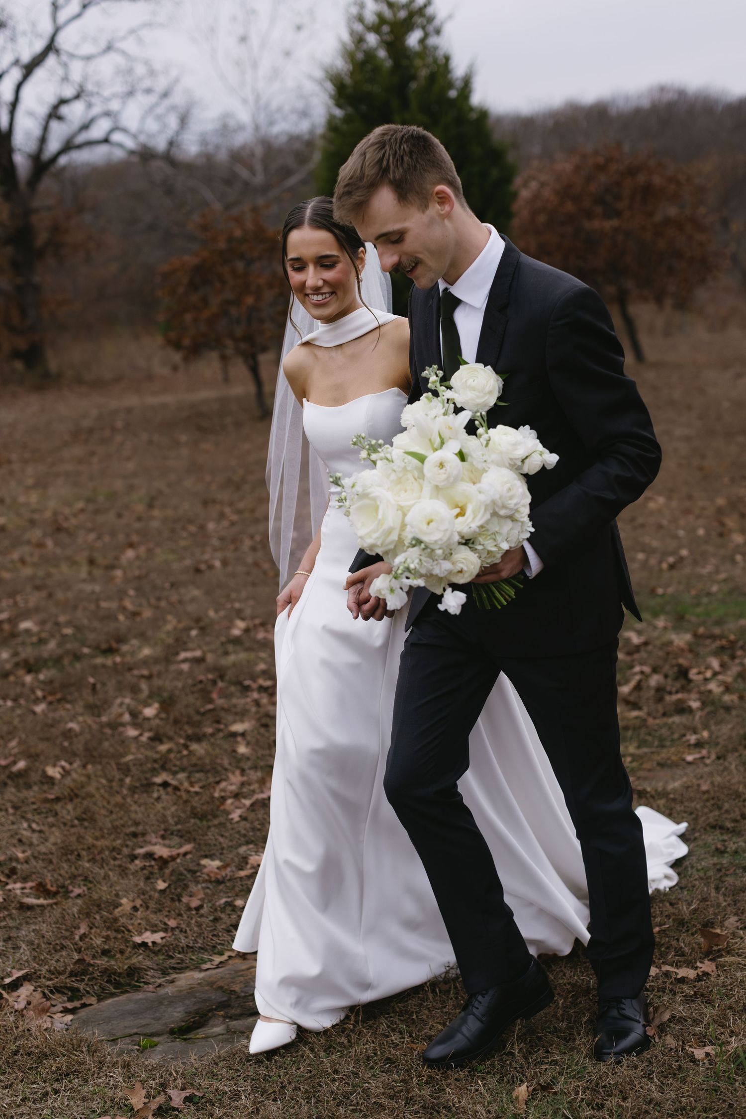 A couple in wedding attire walking together through a winter field holding white flowers.